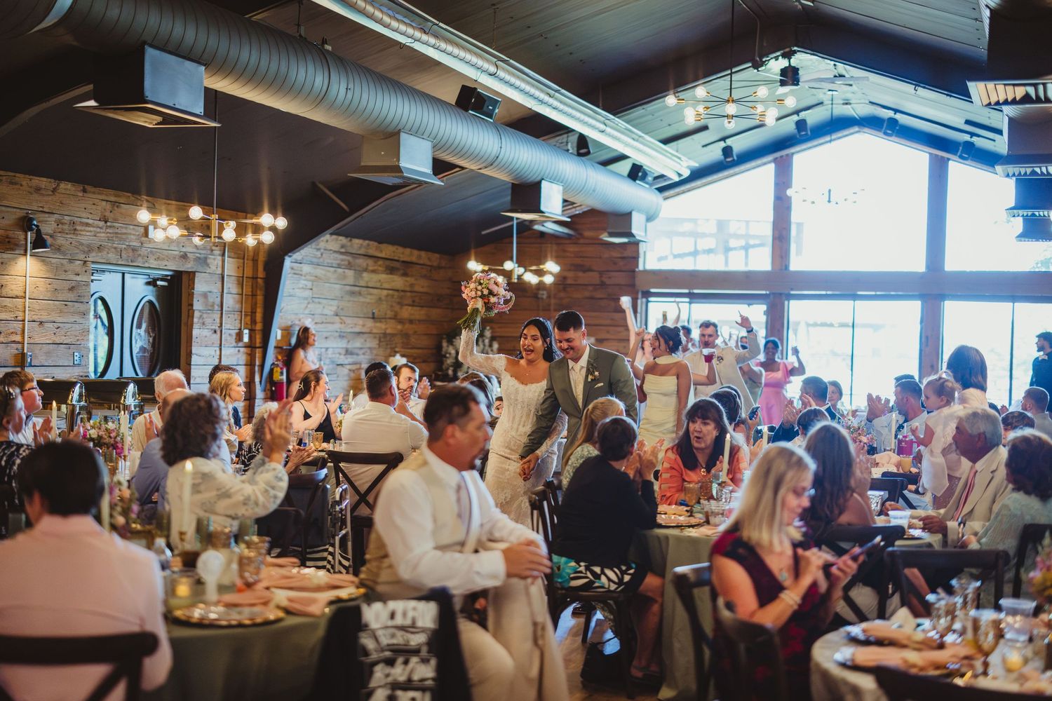 Elegant wedding reception in a rustic brick venue with chandeliers, guests dining at decorated tables.