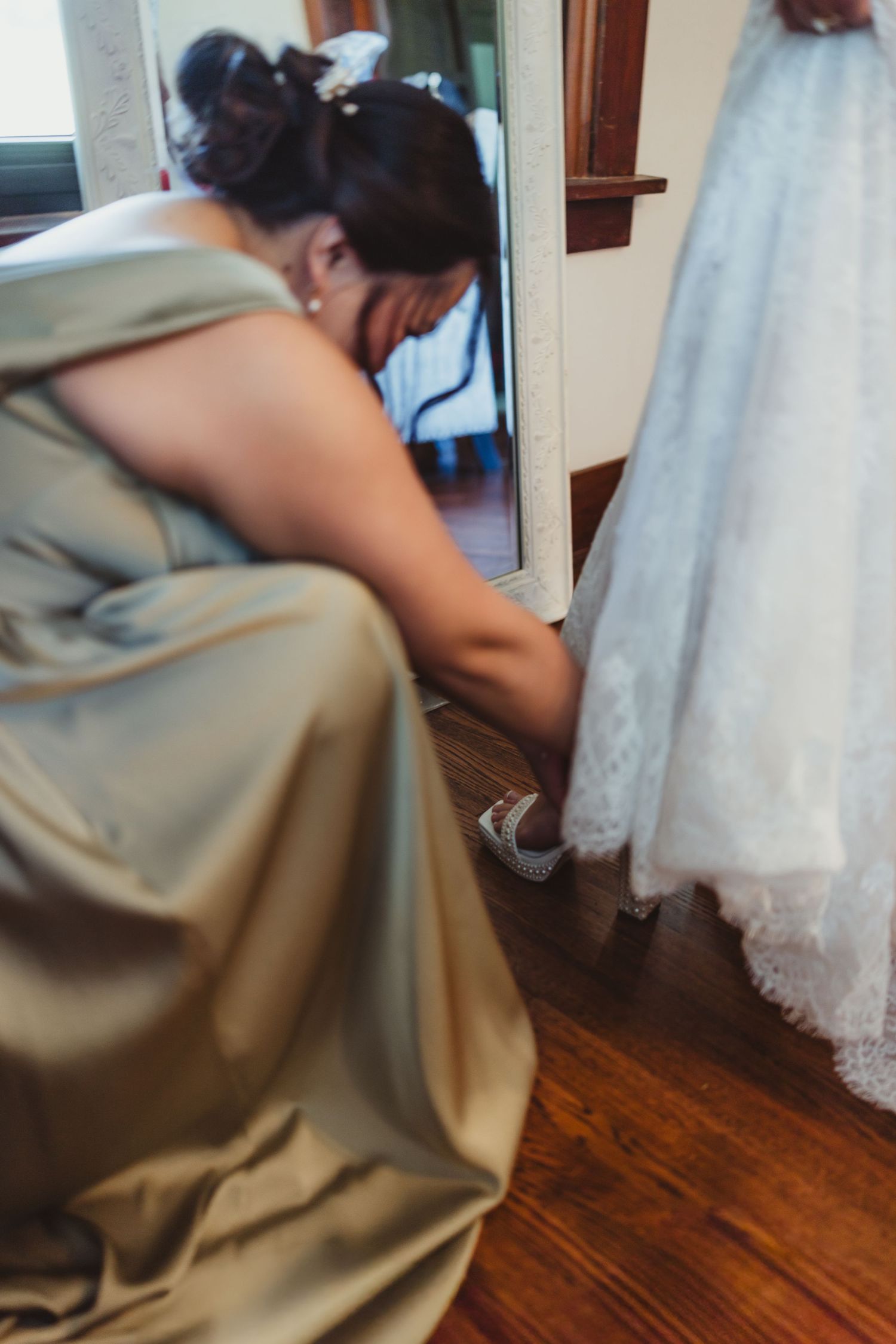 Bridesmaid in gold gown helping bride with her wedding shoes on hardwood floor near mirror.