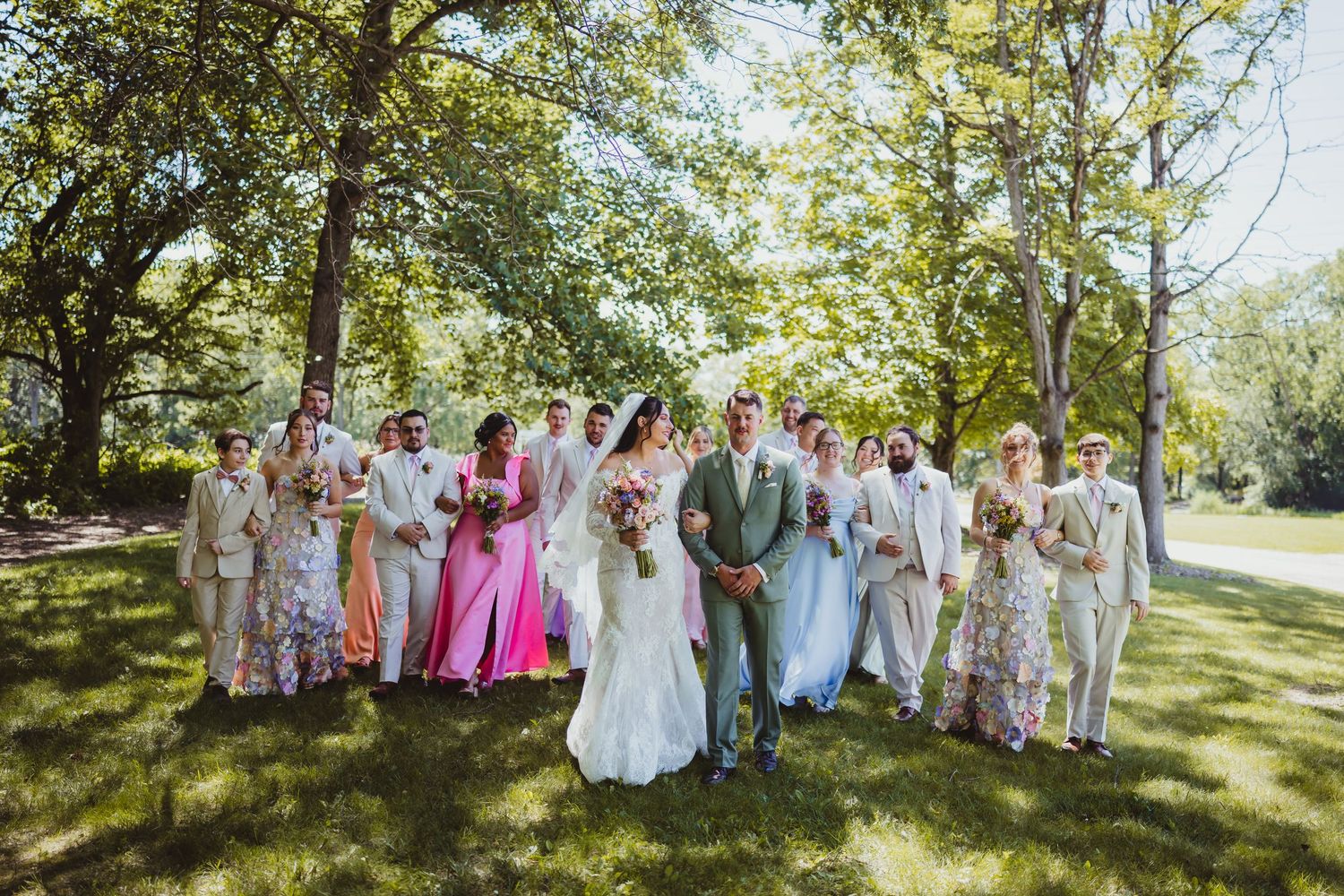 Wedding party walks outdoors on green lawn under trees, bride in white gown, bridesmaids in colorful dresses, groomsmen in tan suits.