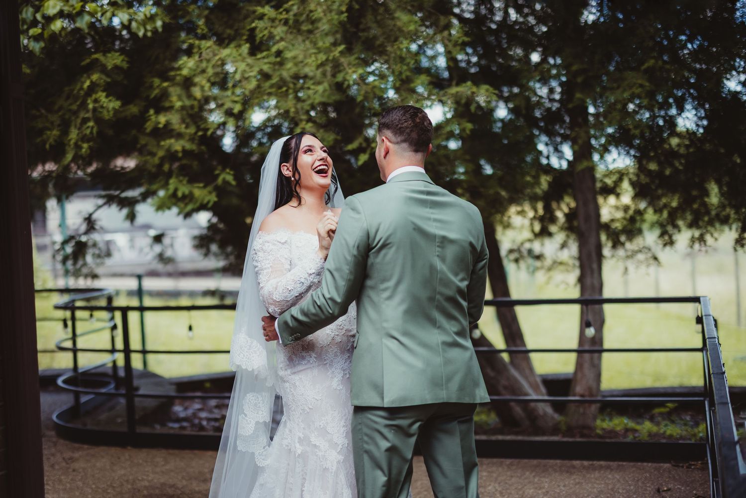 Bride and groom share a joyful moment outdoors, with bride in white gown and veil, groom in sage green suit.