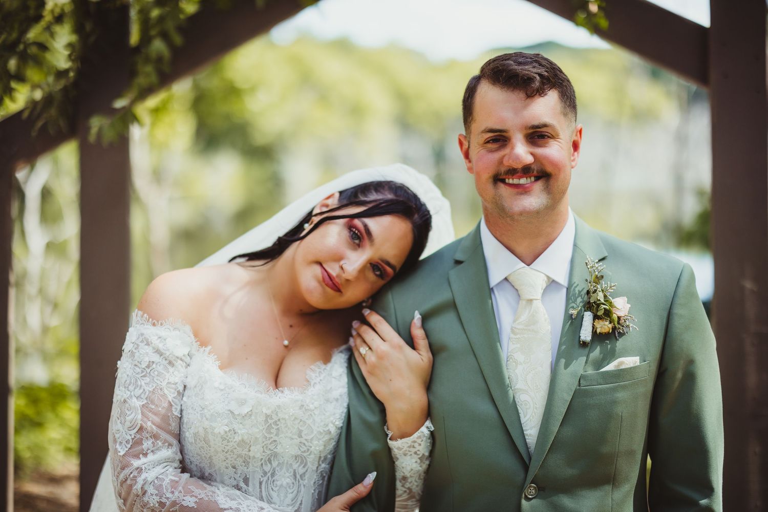 Bride in lace gown leans on groom in sage green suit with boutonniere, posing under wooden arbor outdoors.