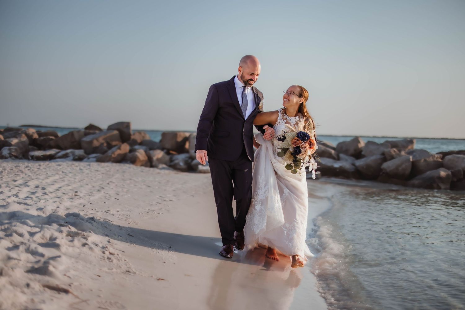A couple walks along a rocky beach at sunset during their wedding photo session.