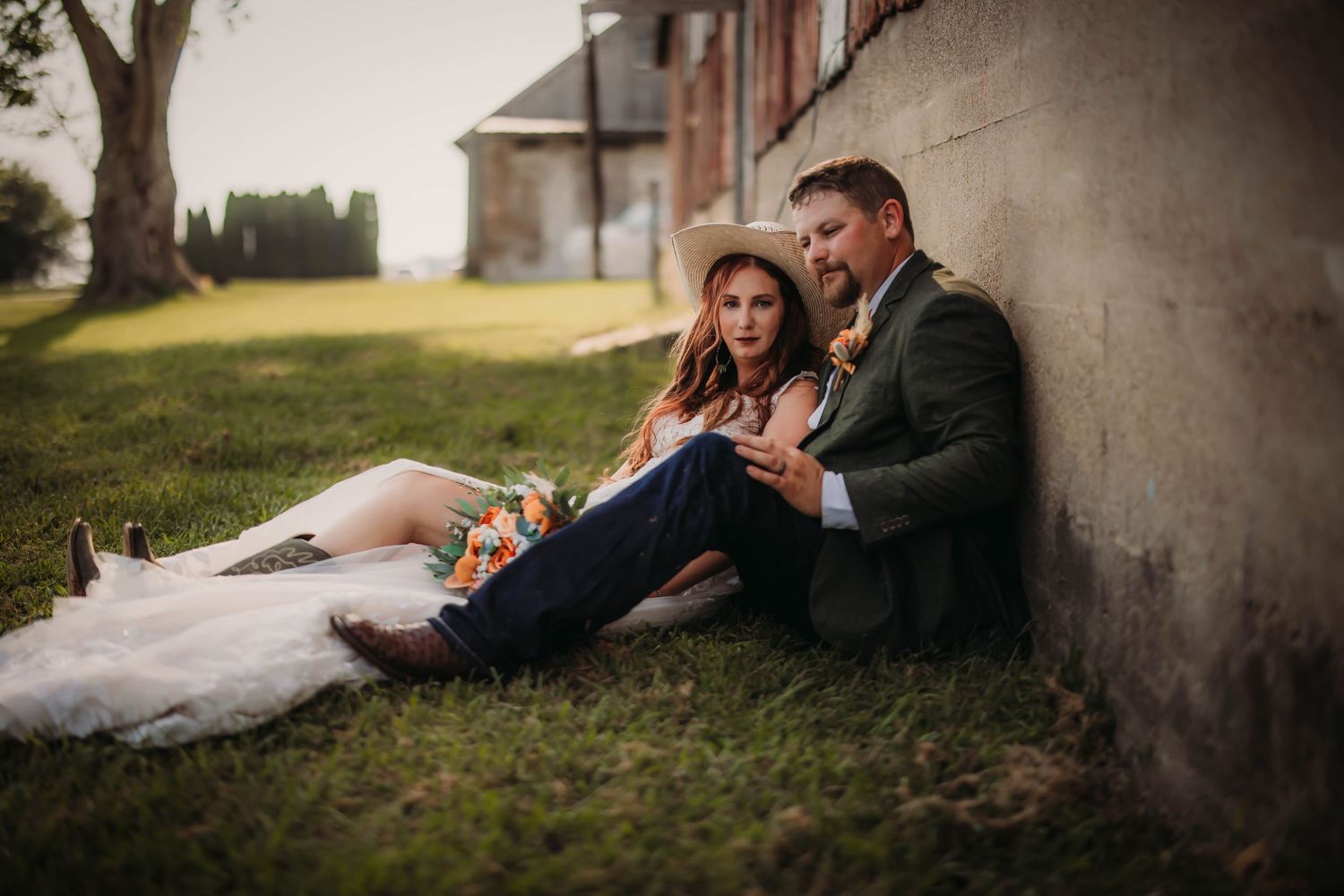 Two people sit on grass against stone wall while embracing in romantic wedding attire.