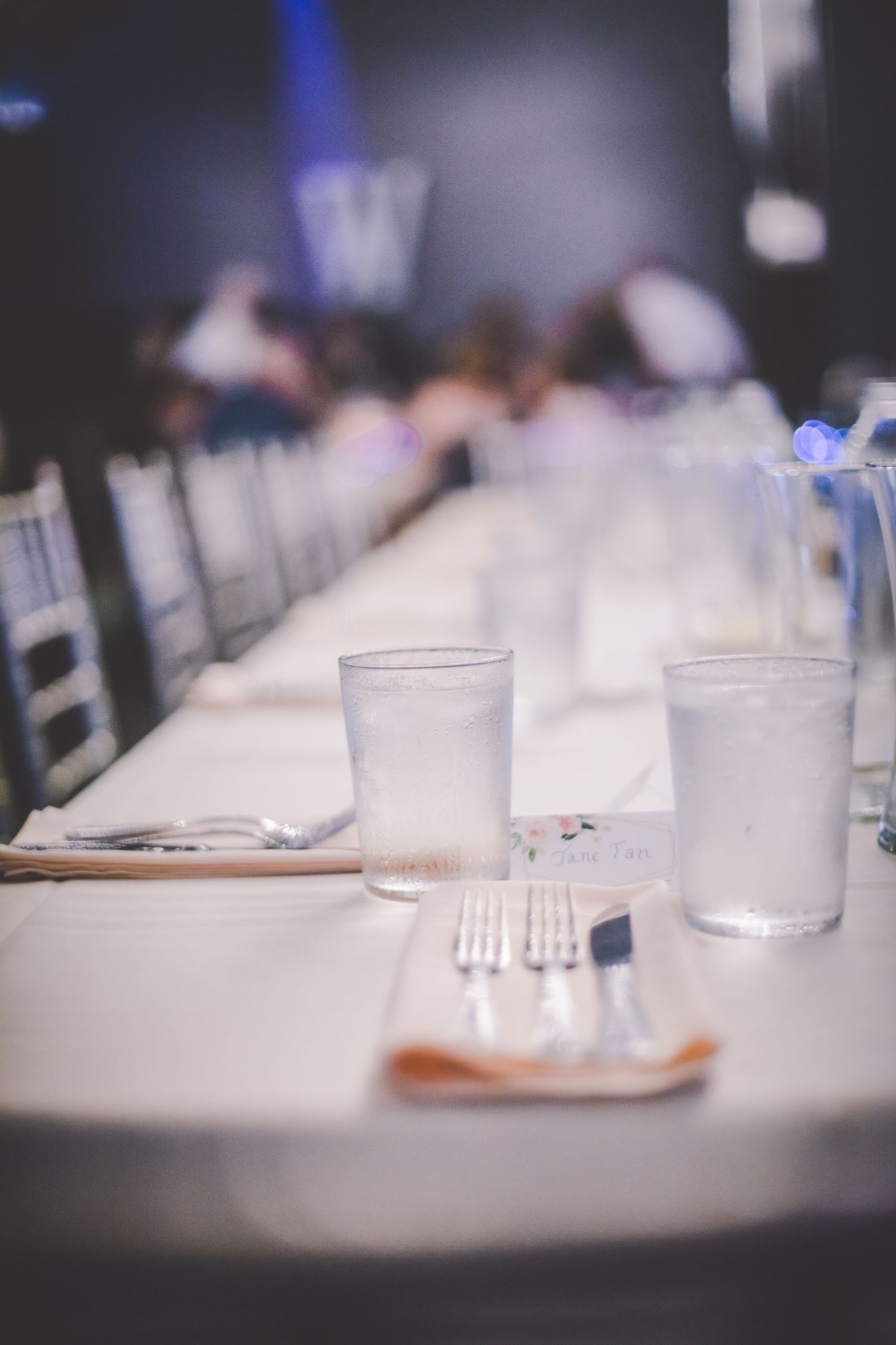 Place settings at a wedding reception with water glasses and silverware on a white tablecloth.
