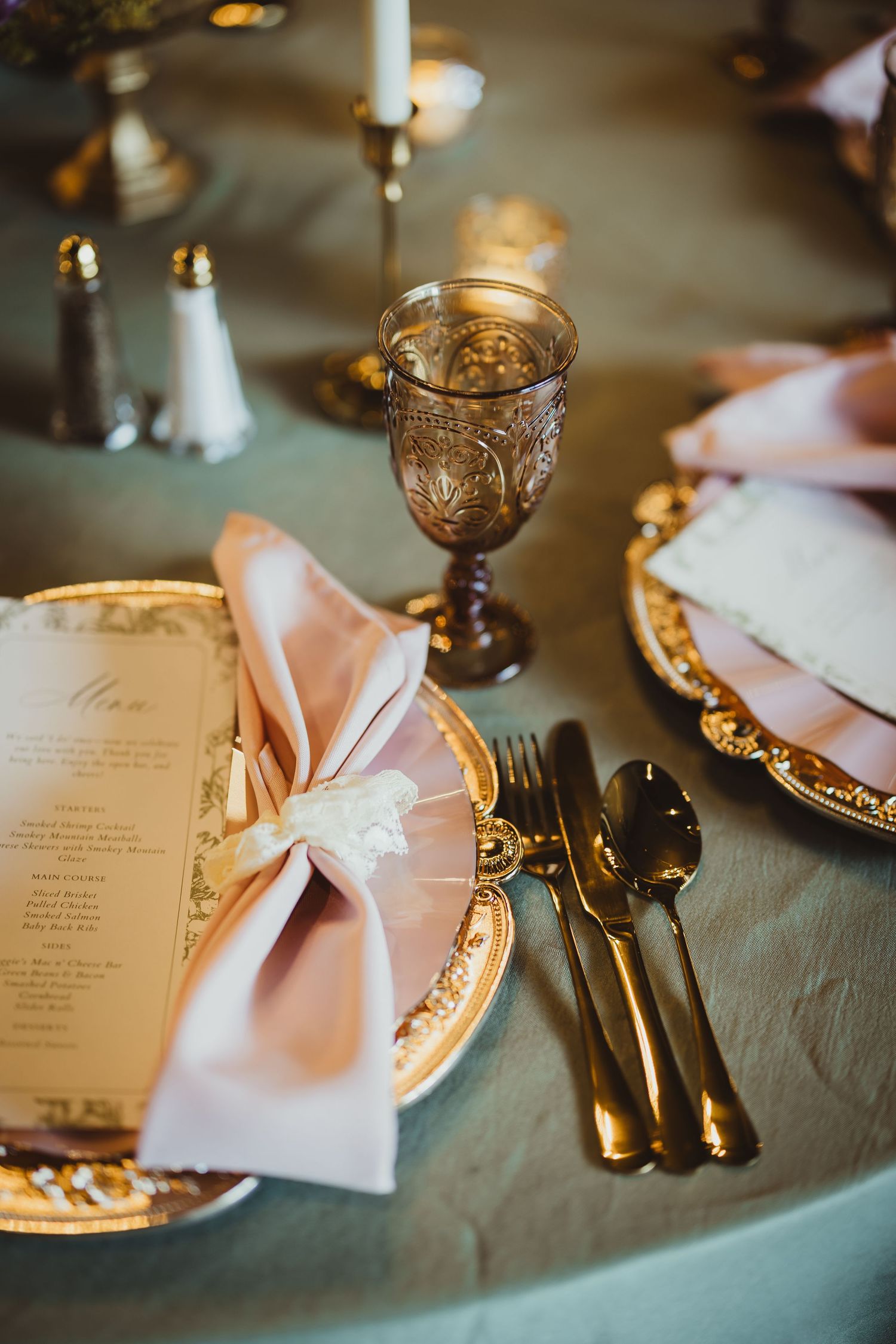 Elegant wedding table setting with gold-rimmed plates, pink napkins, amber goblets, and gold cutlery on a sage green tablecloth.
