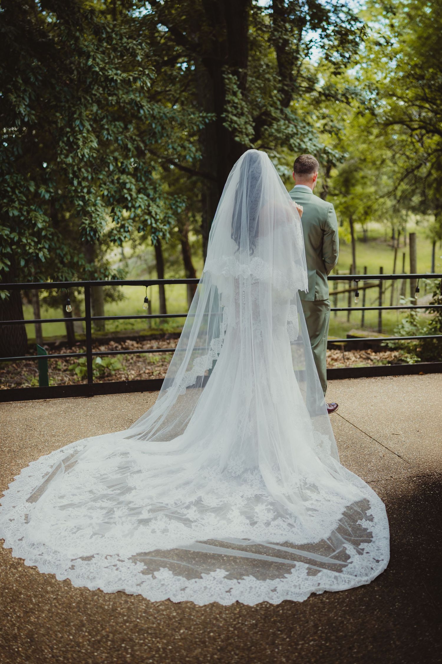 Bride in a long lace-trimmed cathedral veil and groom in sage suit stand together outdoors near a wooden fence.