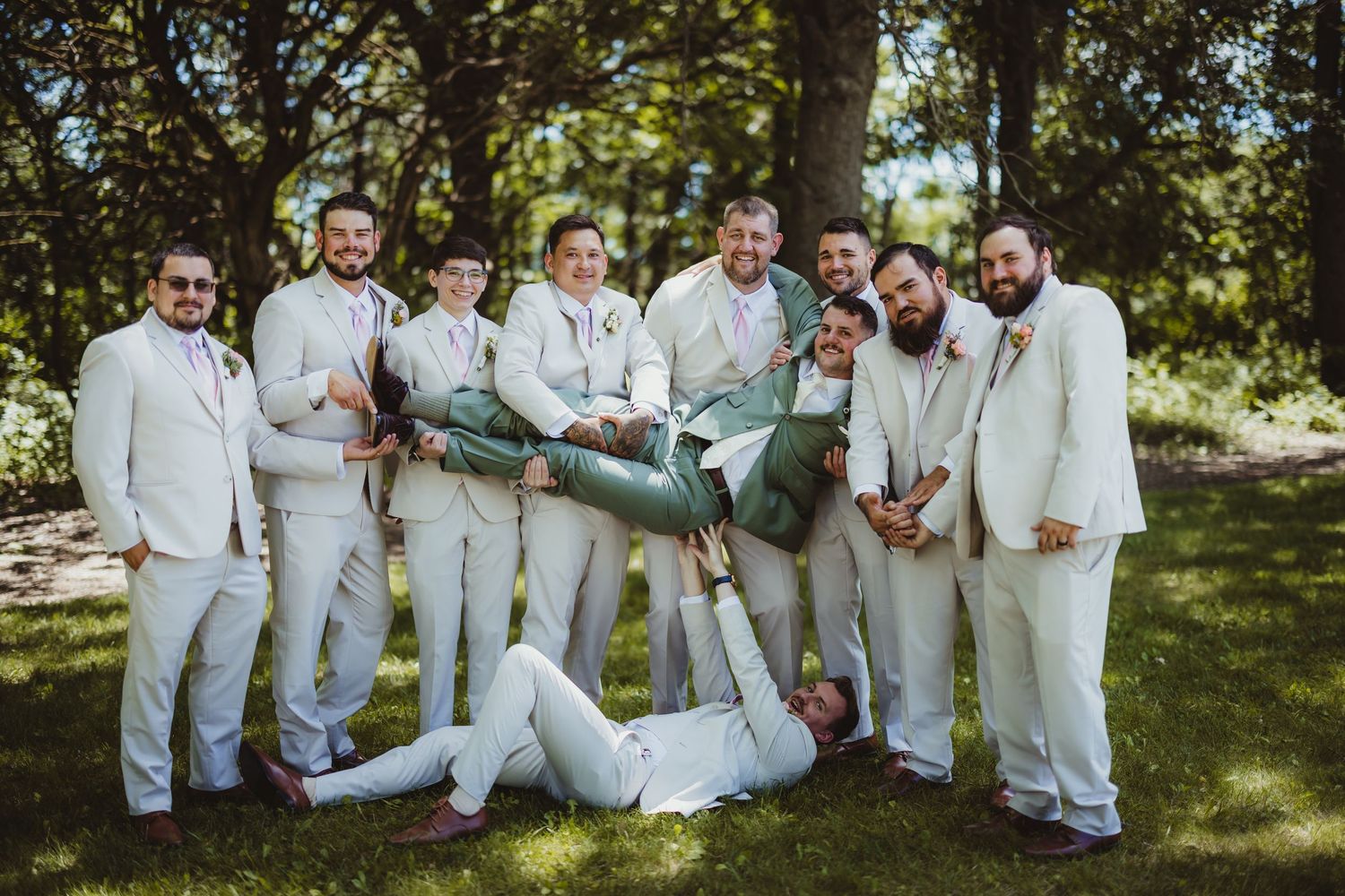 Groomsmen in white suits playfully lifting the groom outdoors at a wedding, surrounded by lush green trees.