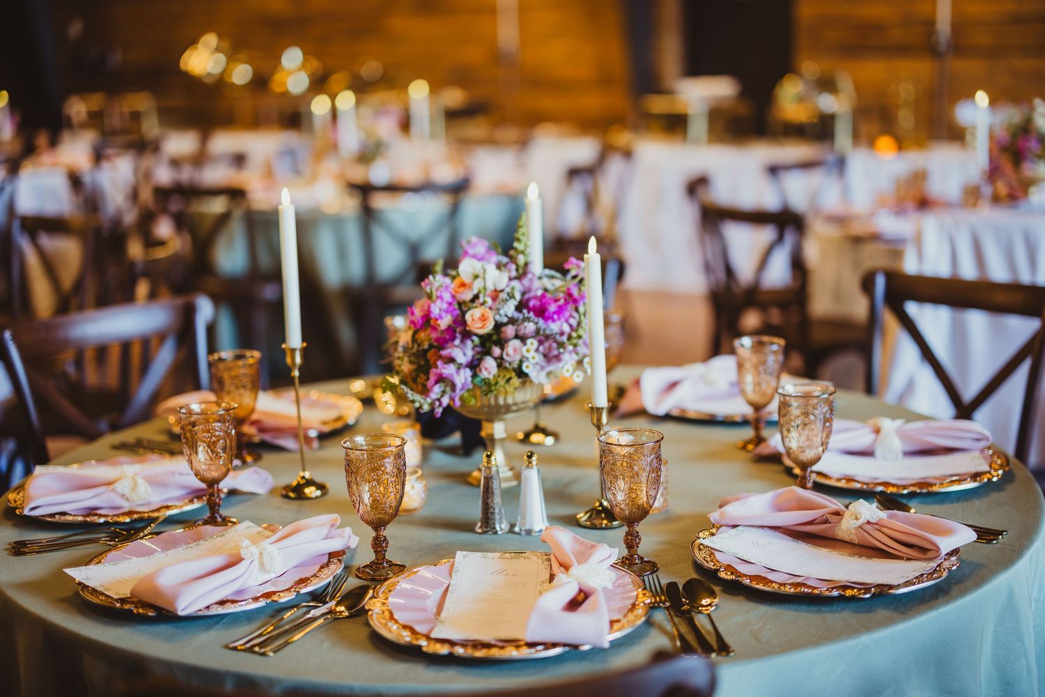 Elegant wedding reception table with gold chargers, pink floral centerpiece, amber glasses, and white candles on sage linen.
