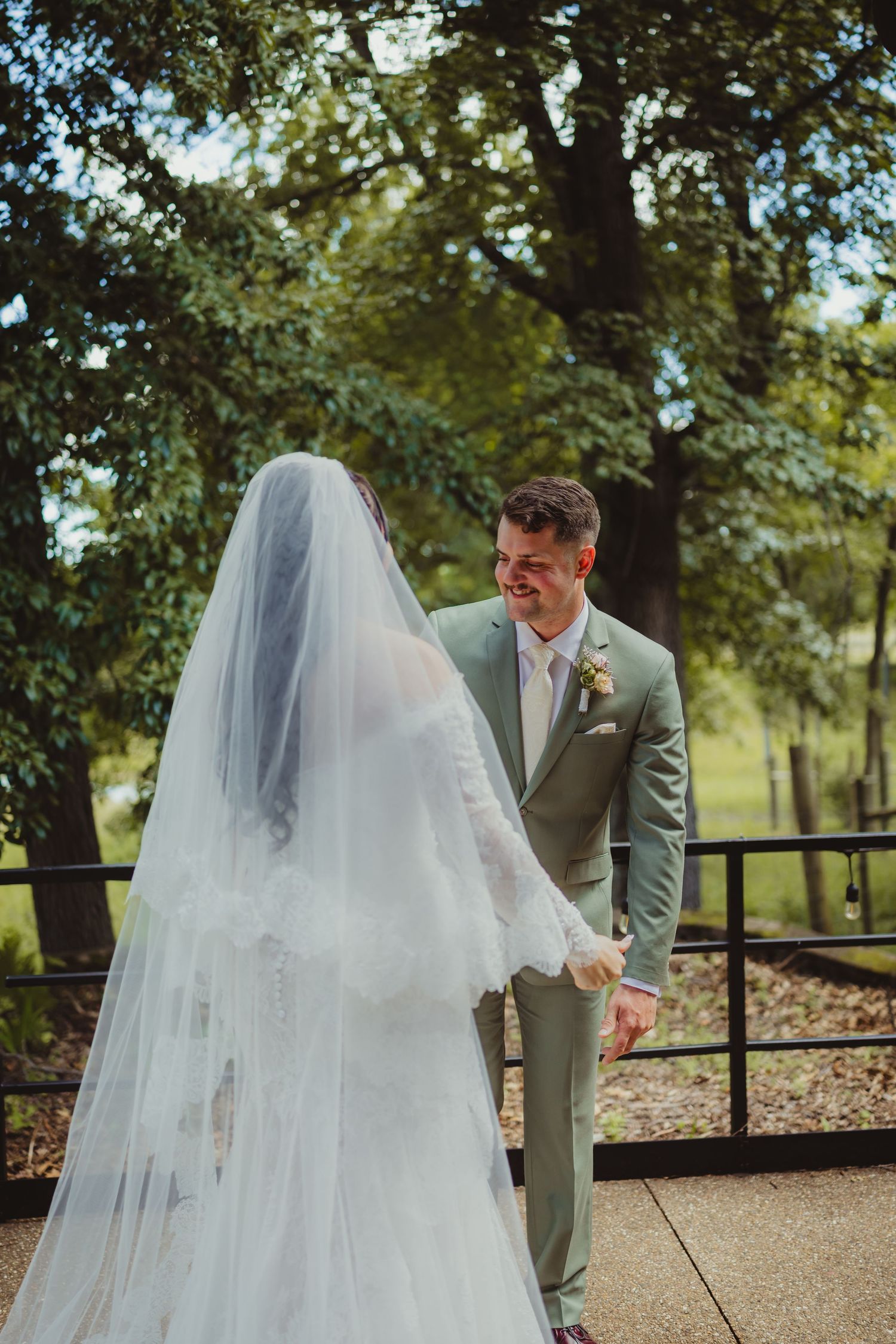 Groom in sage green suit smiling at bride in white lace gown and veil during outdoor wedding first look.