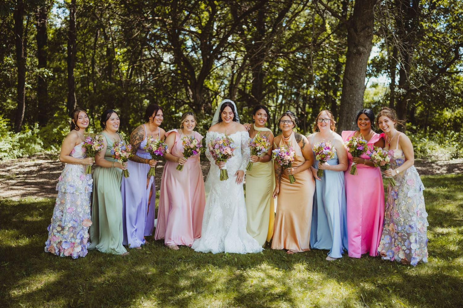 Bride in white gown poses with colorful bridesmaids in rainbow dresses holding bouquets outdoors.