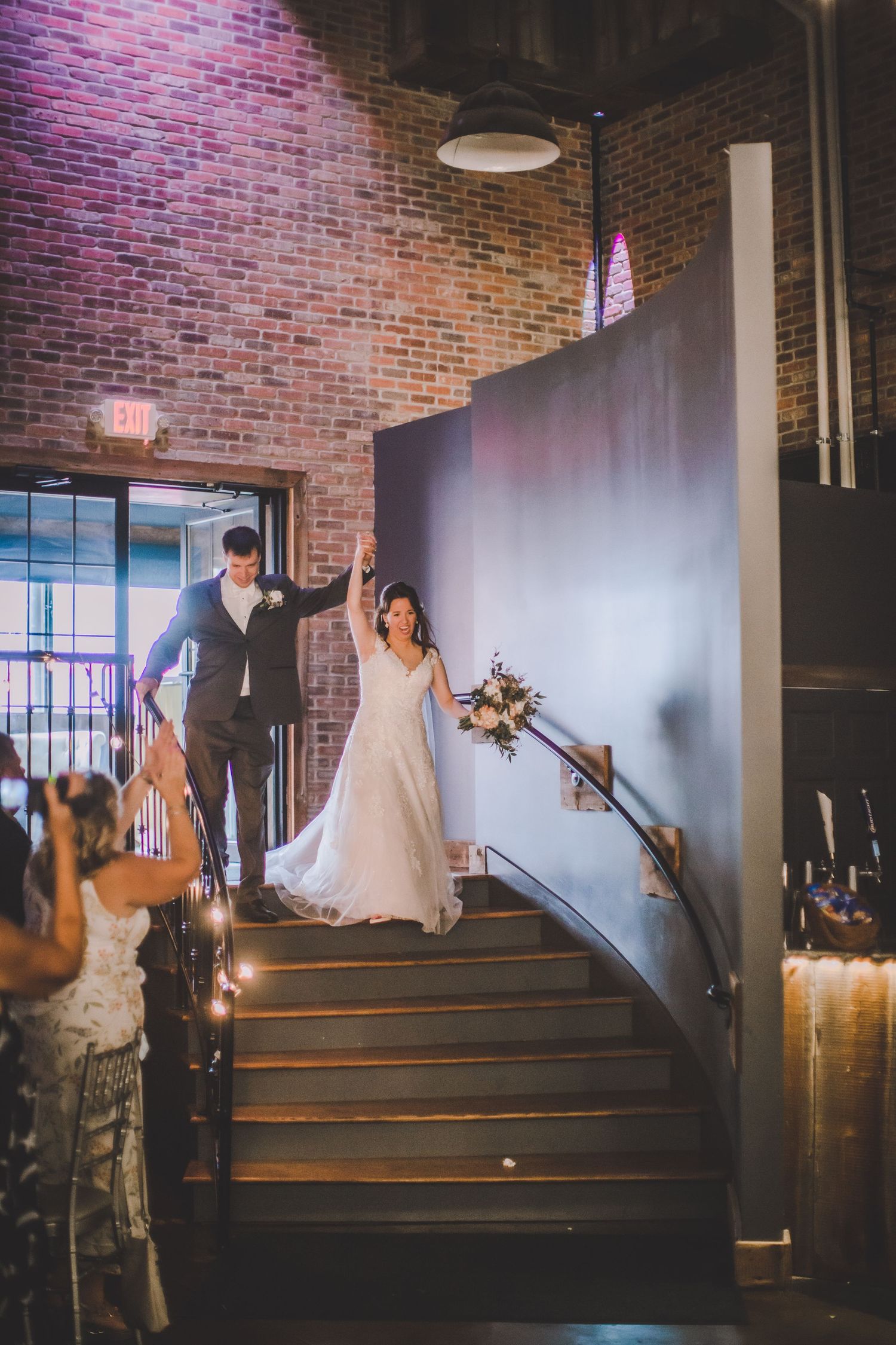 Wedding couple descending brick-walled staircase in industrial-chic venue.
