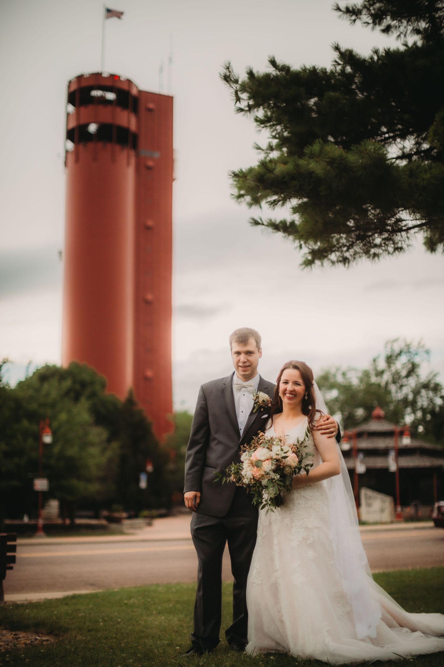 Wedding couple poses in front of tall red tower on grassy lawn.
