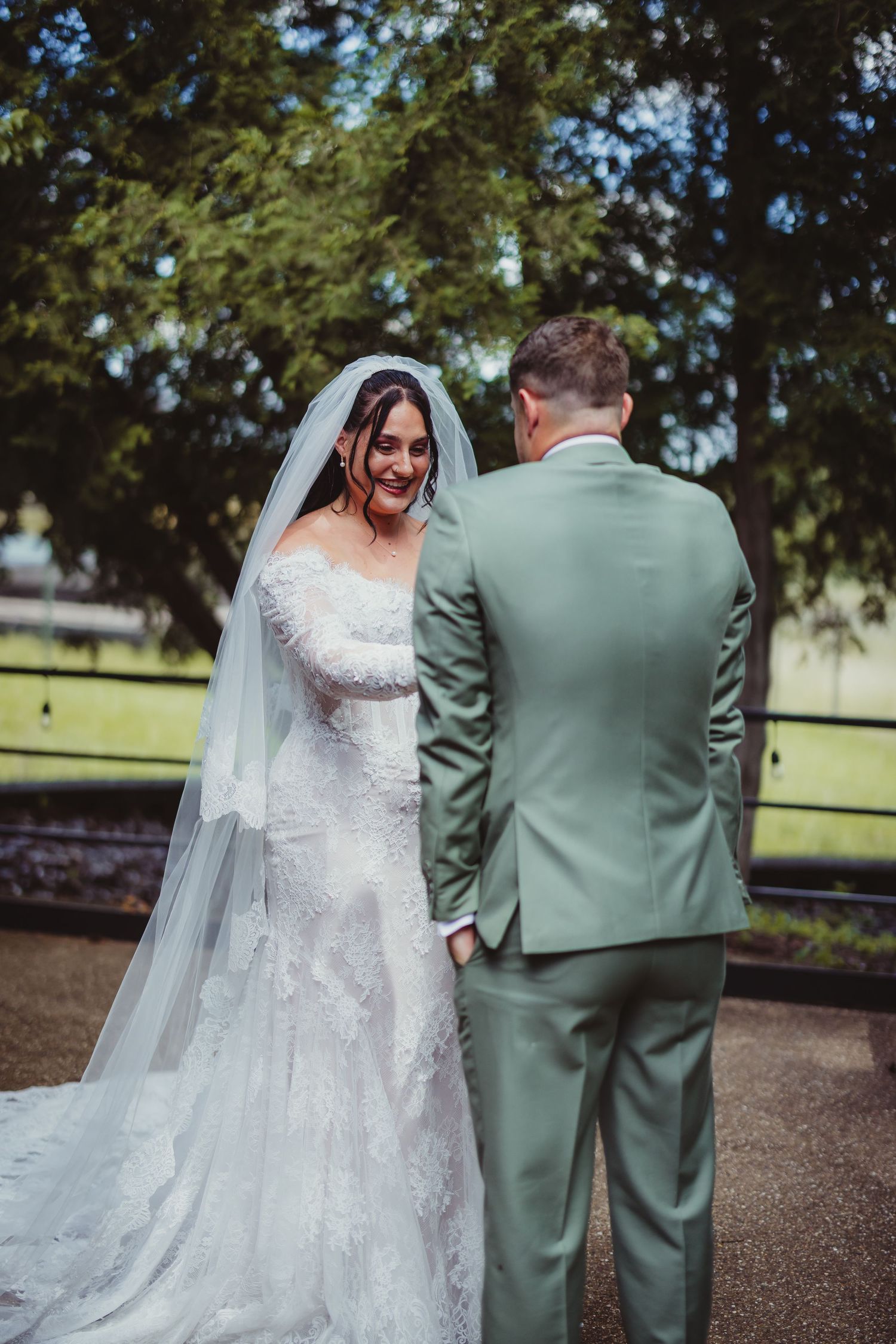 Bride in lace gown and veil smiling at groom in sage green suit during outdoor wedding portrait.