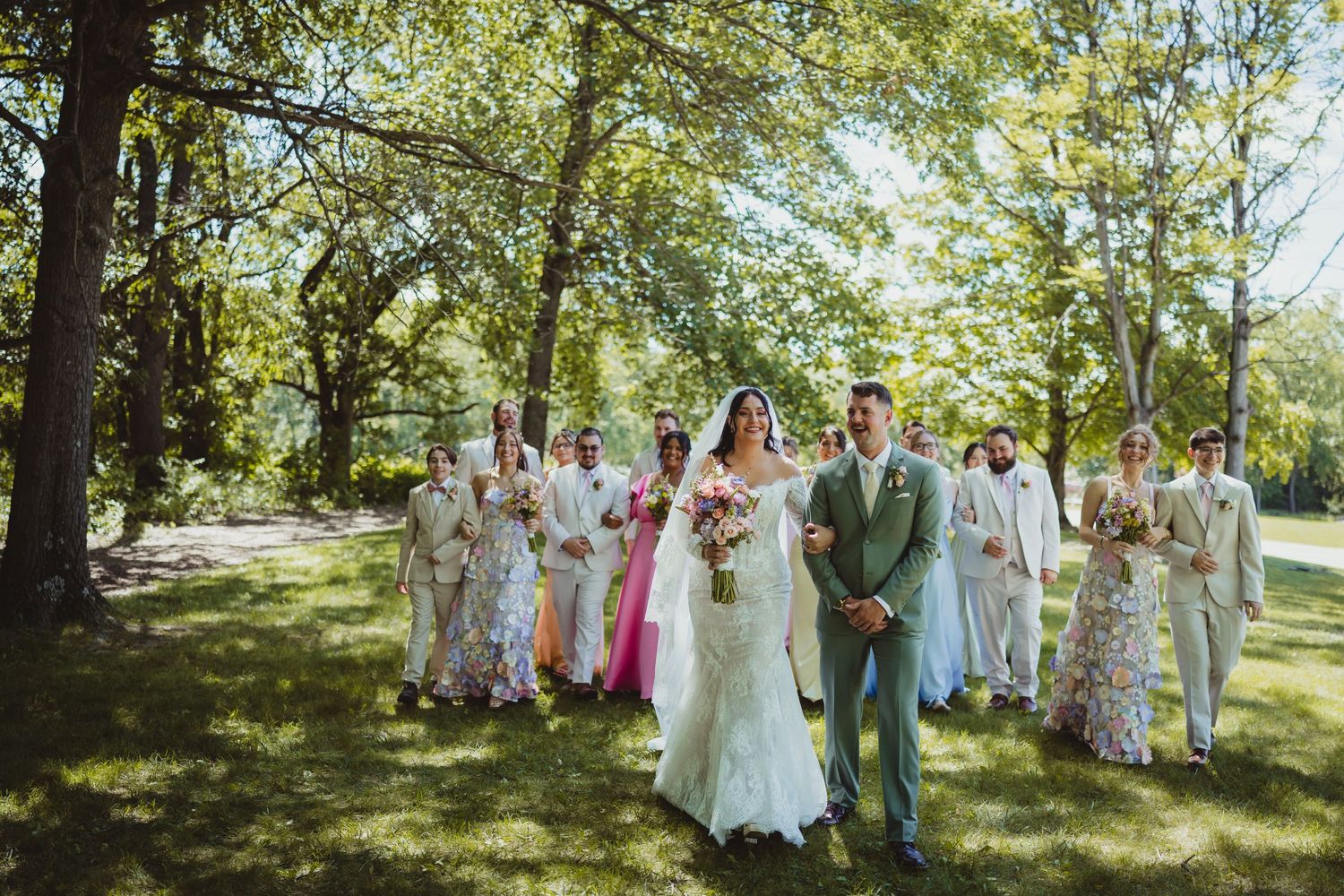 Bride and groom lead wedding party across lush green lawn under tall trees on sunny summer wedding day.