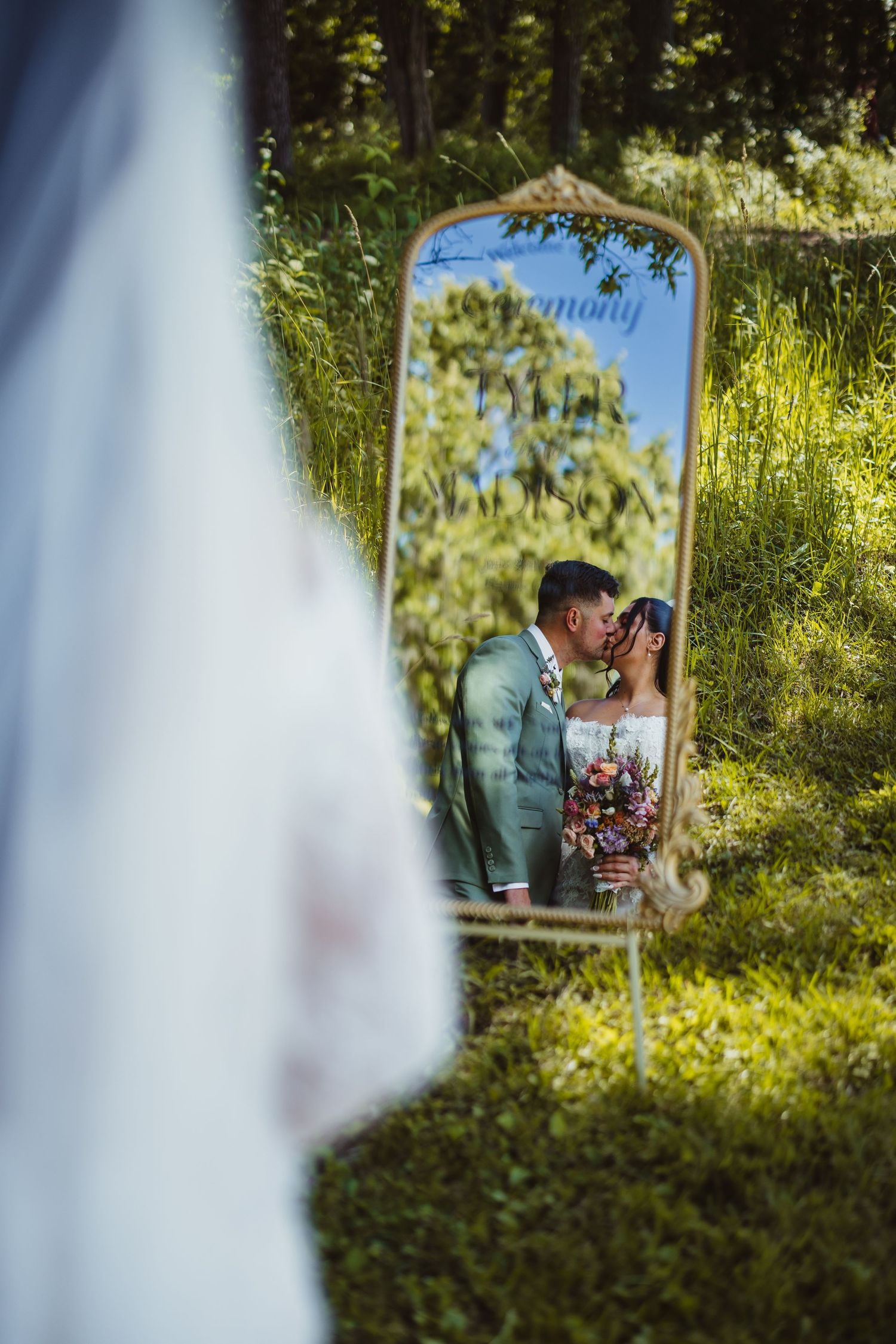 Wedding couple kissing reflected in ornate mirror surrounded by lush green grass outdoors.