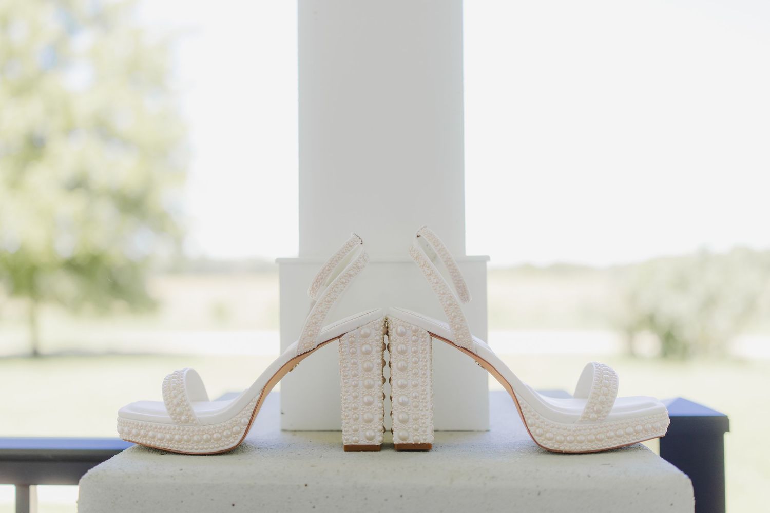 Elegant white bridal heels with pearl embellishments displayed on a ledge with a green outdoor backdrop.