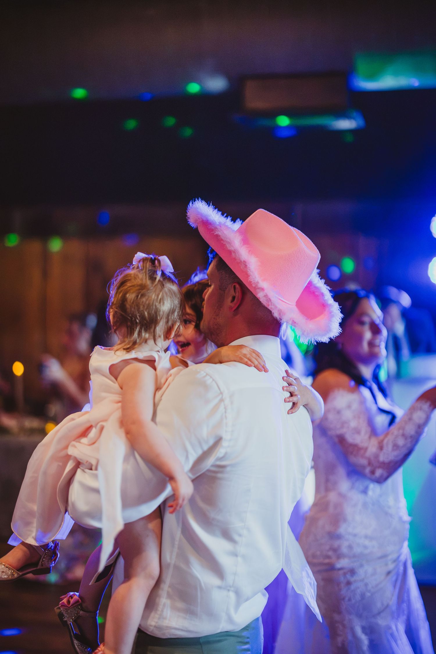 Man in pink cowboy hat holds young girl while dancing at a festive wedding reception with colorful lights.