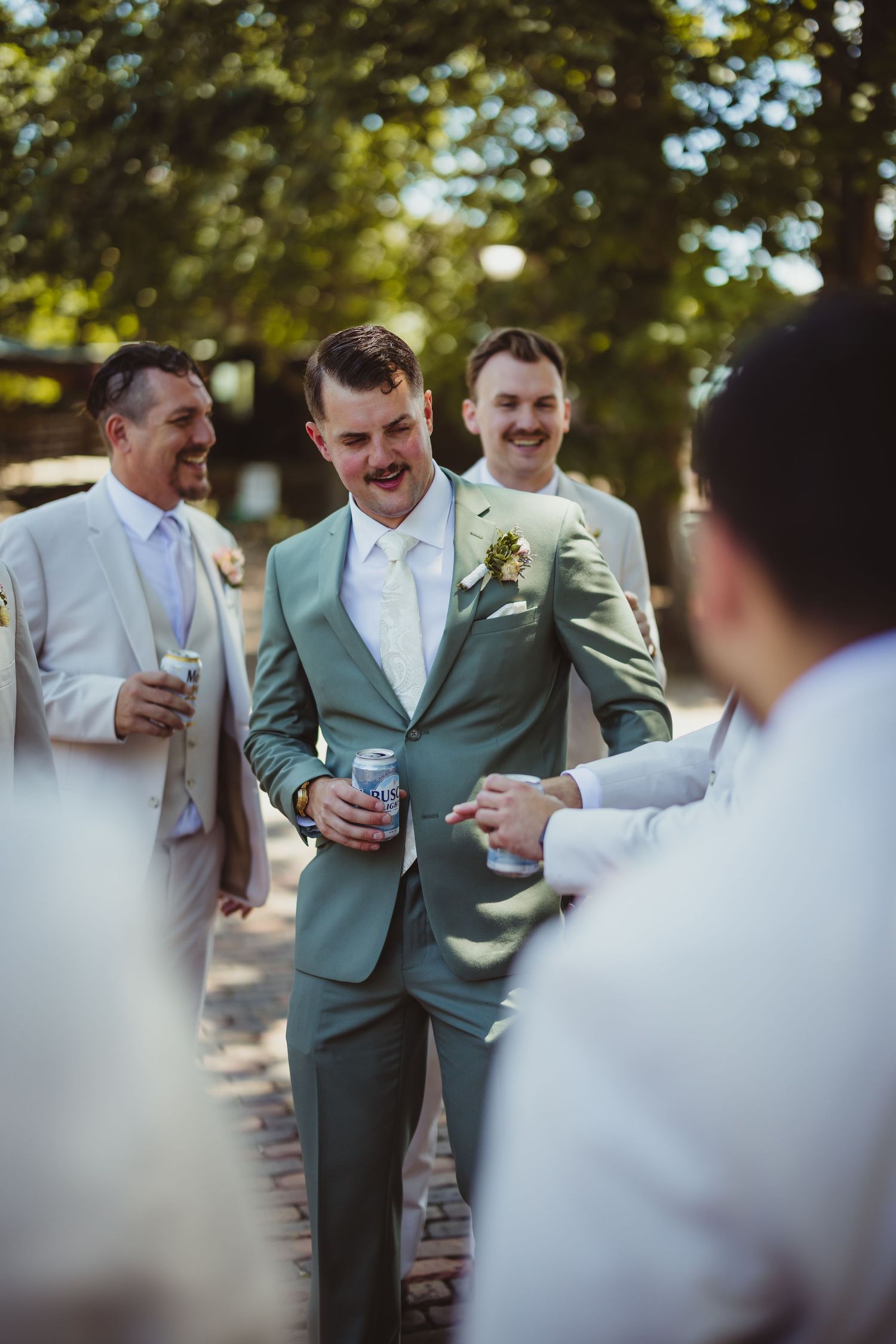 Groom in sage green suit with boutonniere laughing with groomsmen in white suits at outdoor wedding celebration.