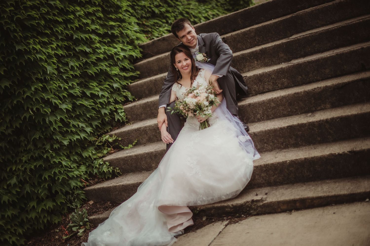 Wedding couple poses playfully on historic stone steps with climbing ivy wall.
