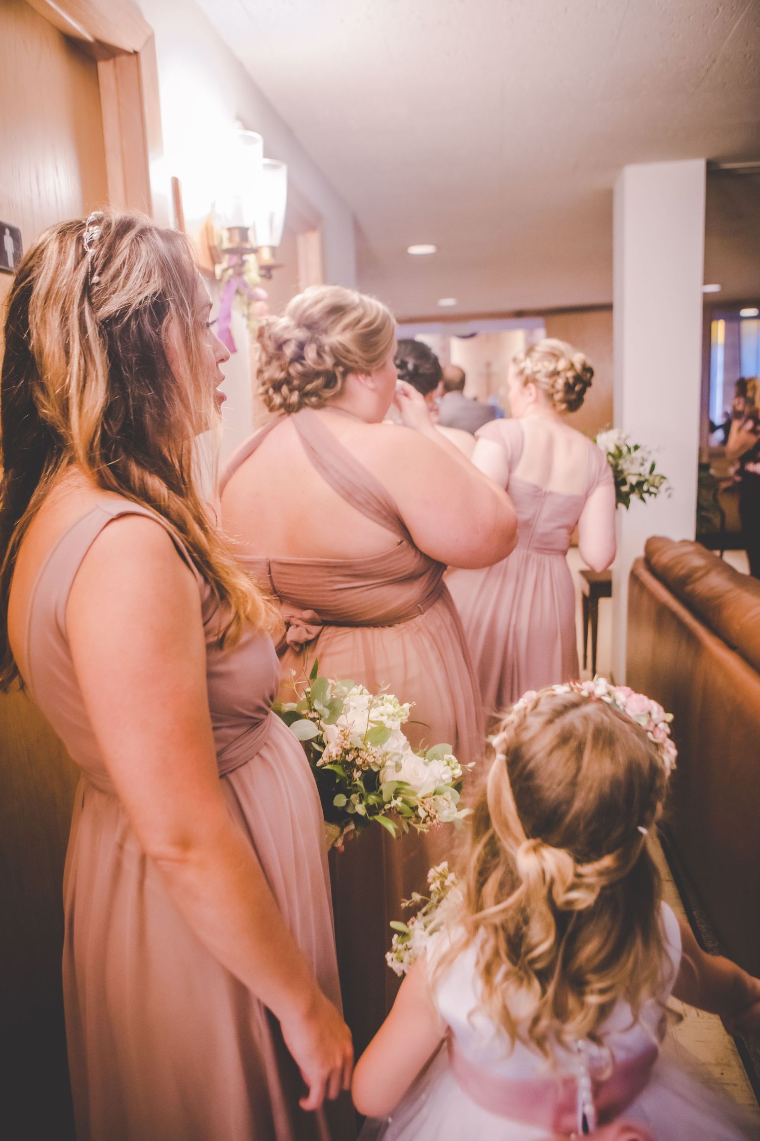 Bridesmaids in blush gowns wait with flower girl in hotel corridor.