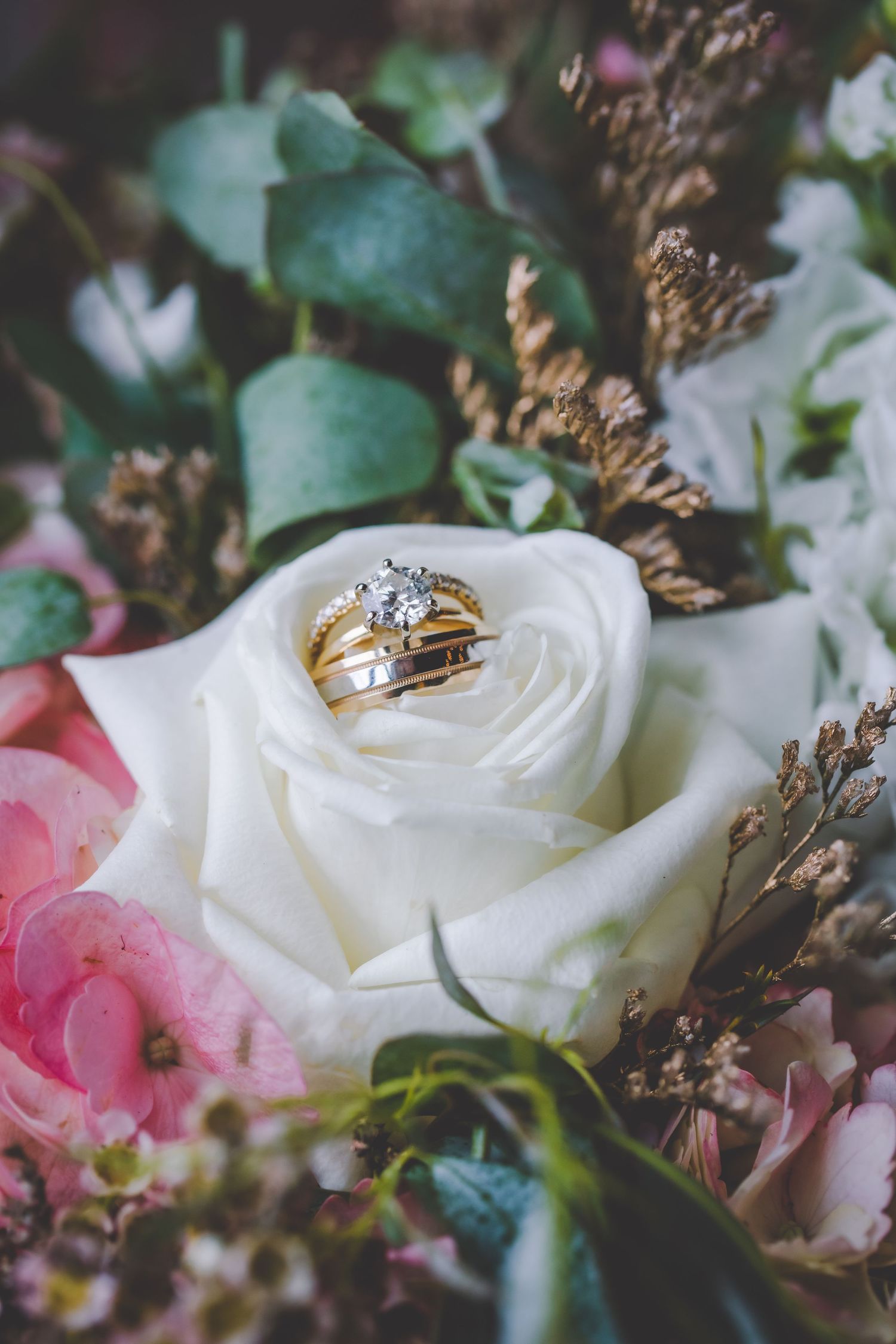 Gold wedding rings displayed on a white rose surrounded by pink flowers and greenery.