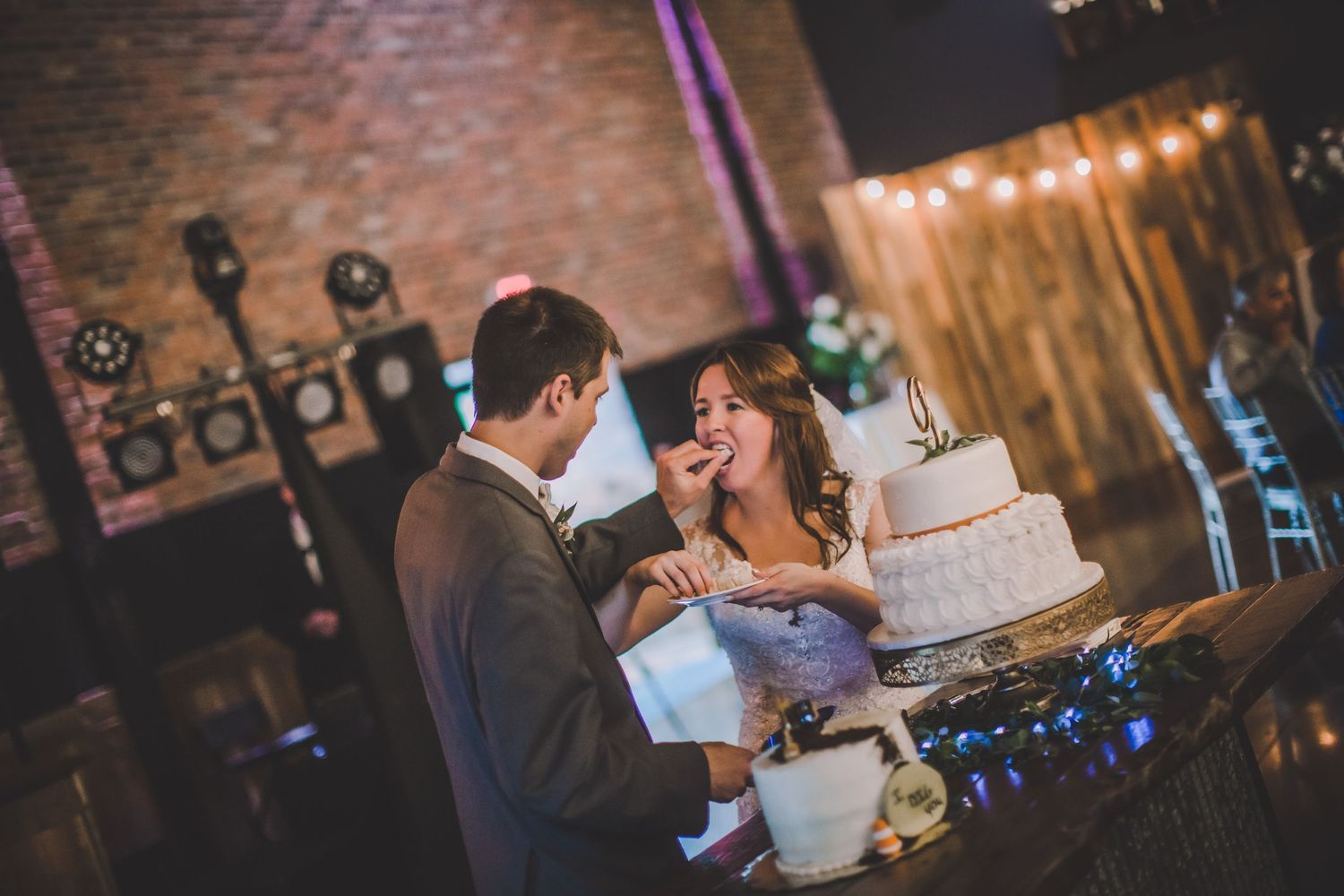 Newlyweds feed each other wedding cake during their reception in a rustic venue with string lights.