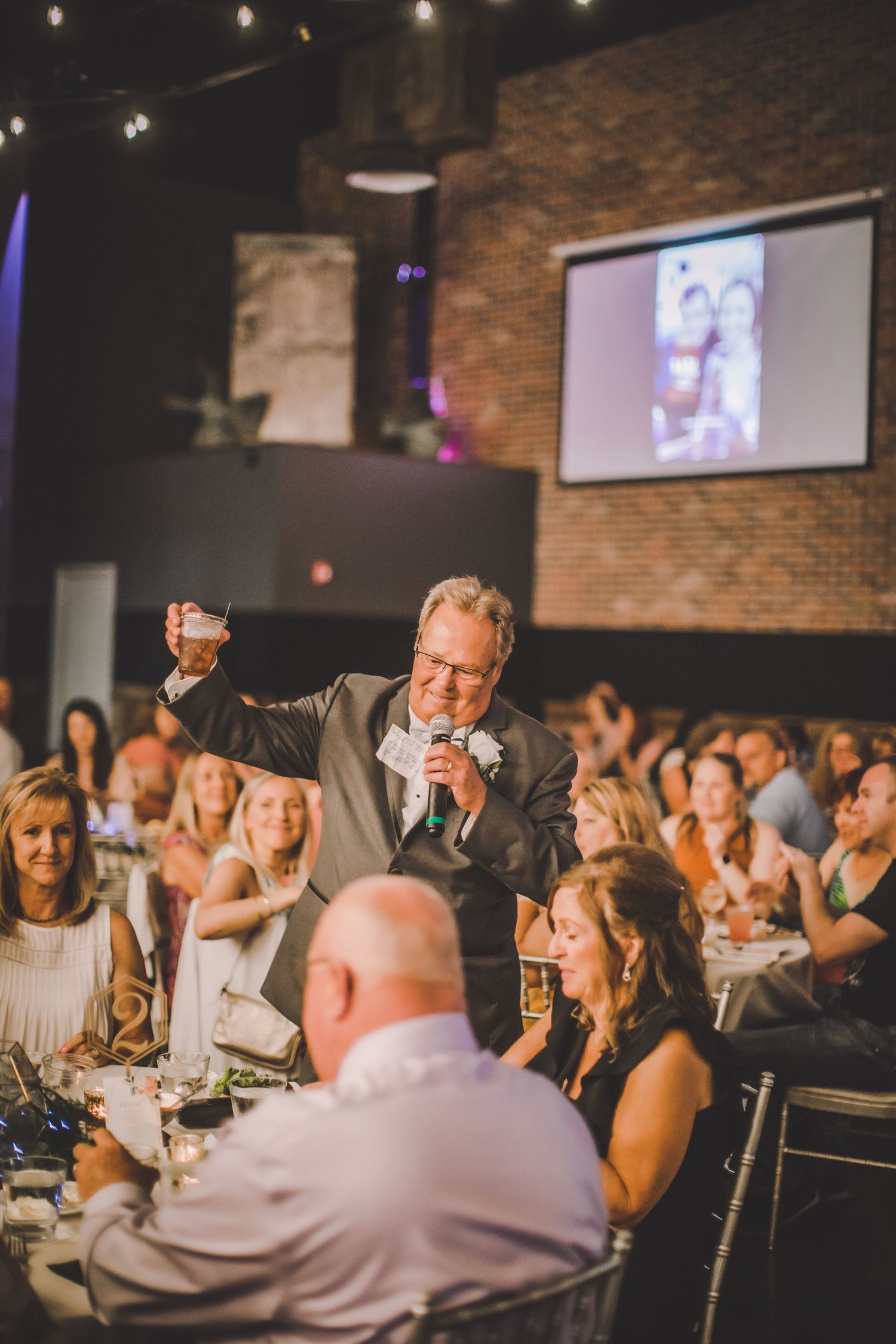 A person raises a glass while giving an animated wedding toast to a room full of guests.