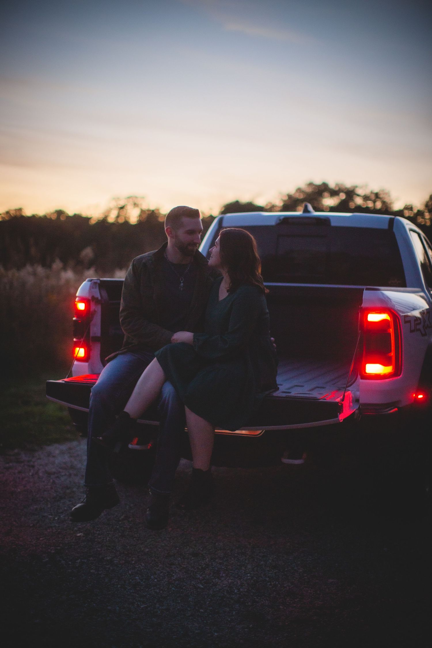 Couple sitting on pickup truck tailgate during sunset with red taillights glowing.
