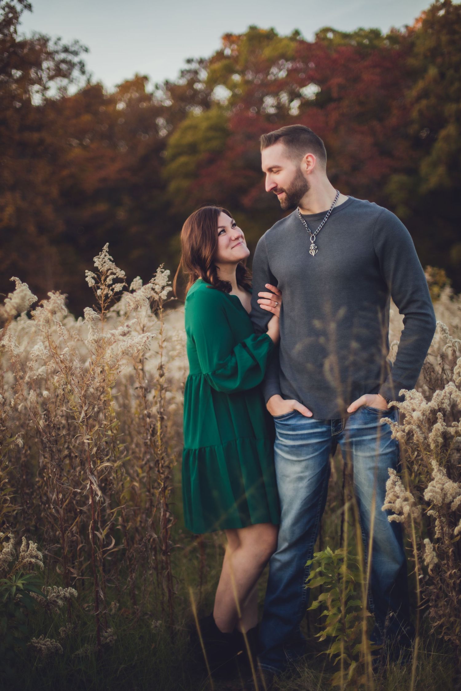 A couple poses together in a field of tall grass, with her in a green dress and him in casual wear.