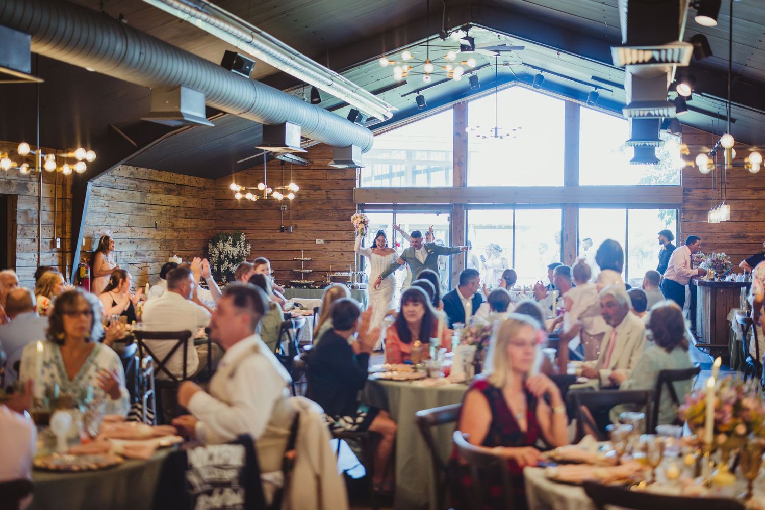 Guests dining at elegant wedding reception in rustic venue with exposed brick, chandeliers, and large windows.