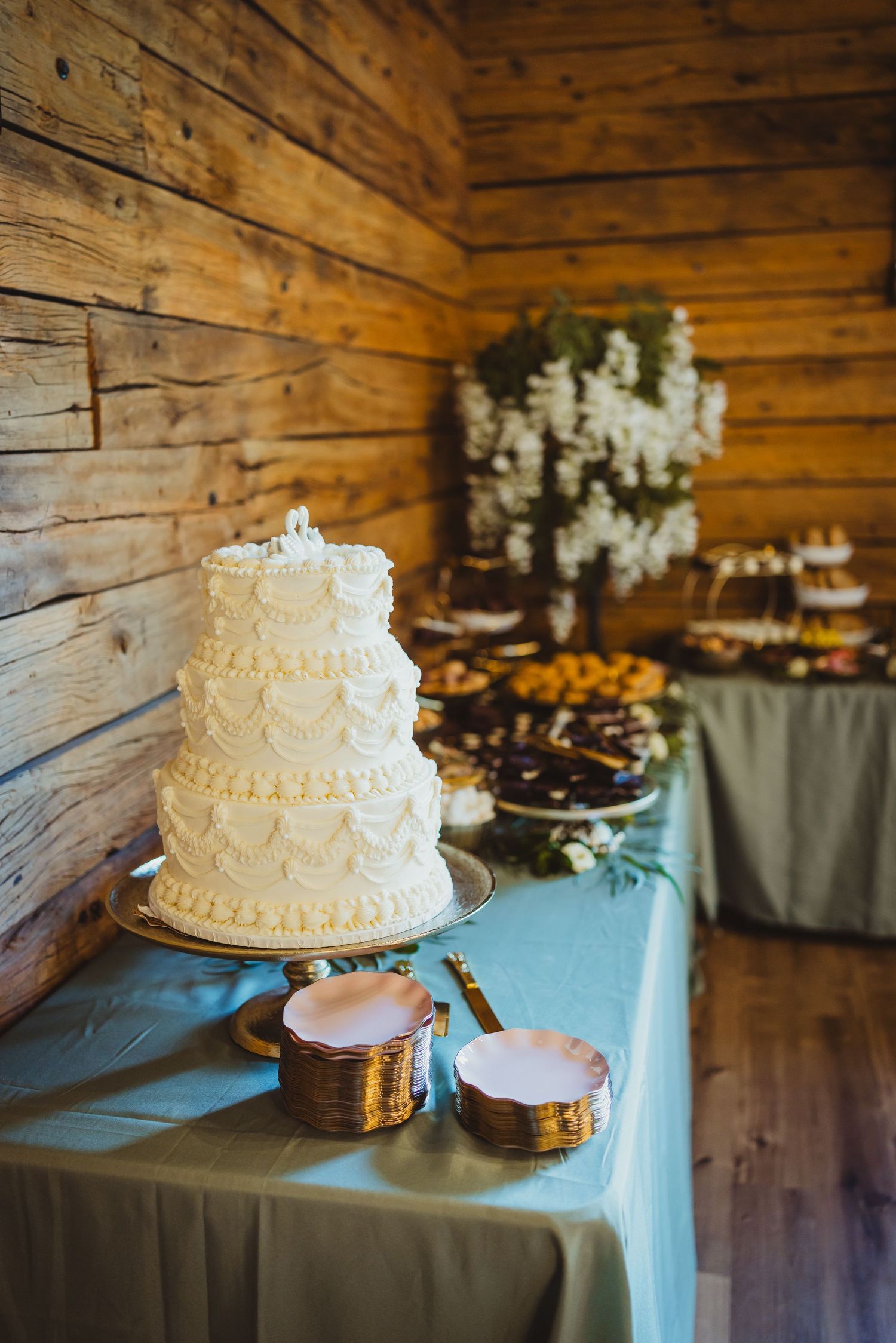 Elegant three-tier white wedding cake on a dessert table with blue tablecloth in a rustic wooden venue.