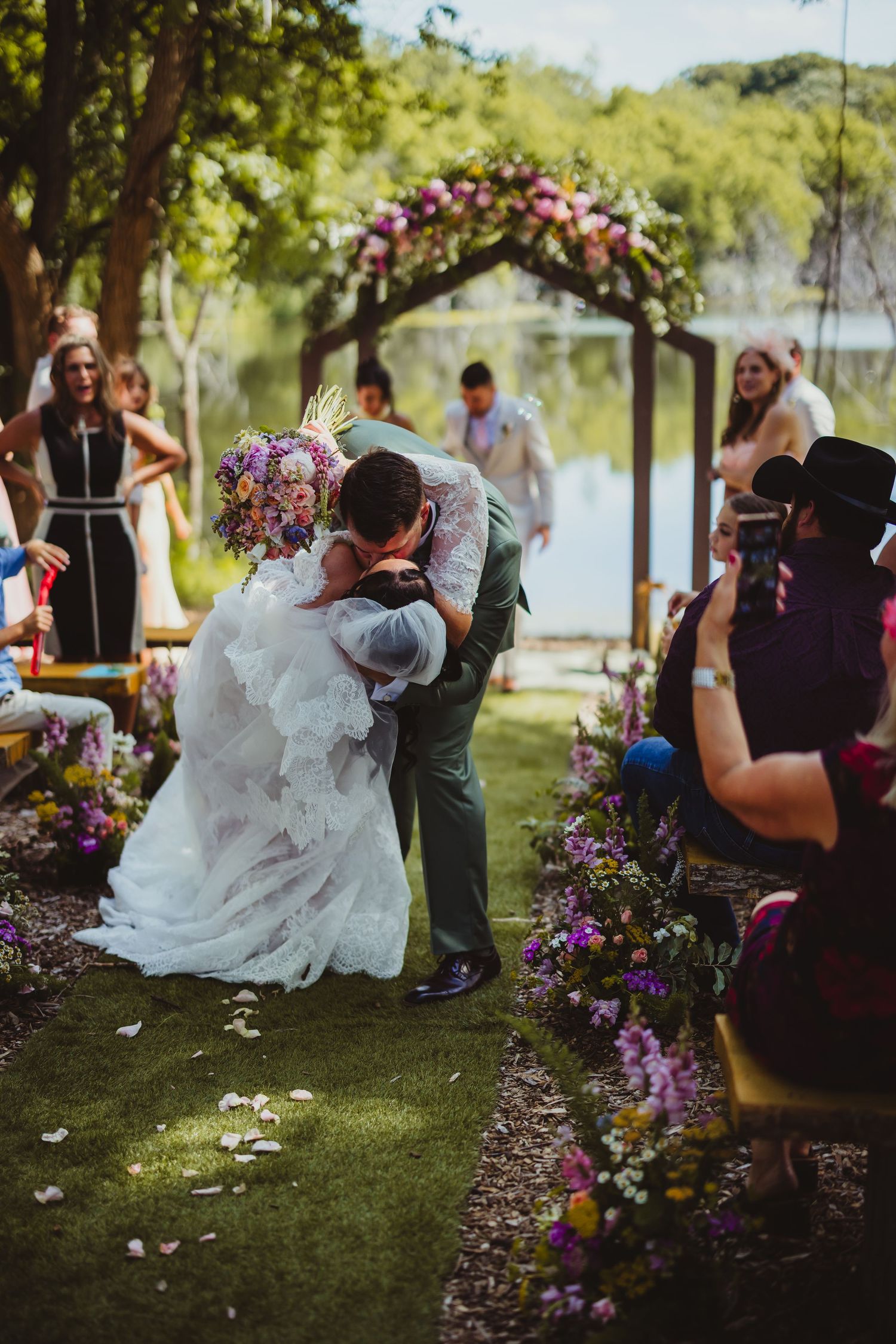 Bride and groom share first kiss at outdoor wedding ceremony with floral arch and colorful blooms lining the aisle.