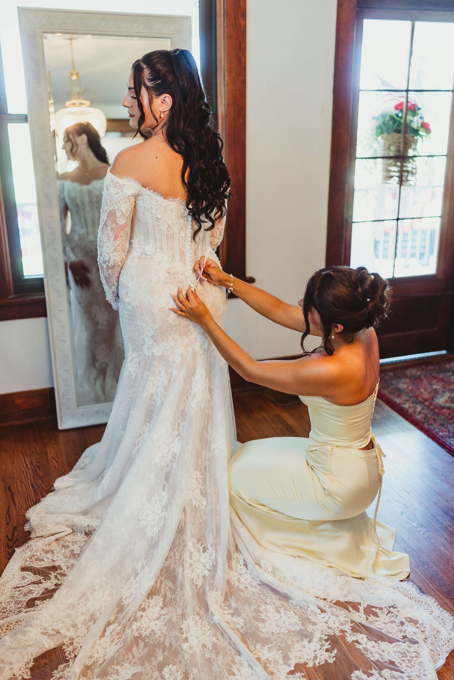 Bridesmaid in yellow dress kneeling to fasten buttons on bride's lace off-shoulder wedding gown in elegant room.
