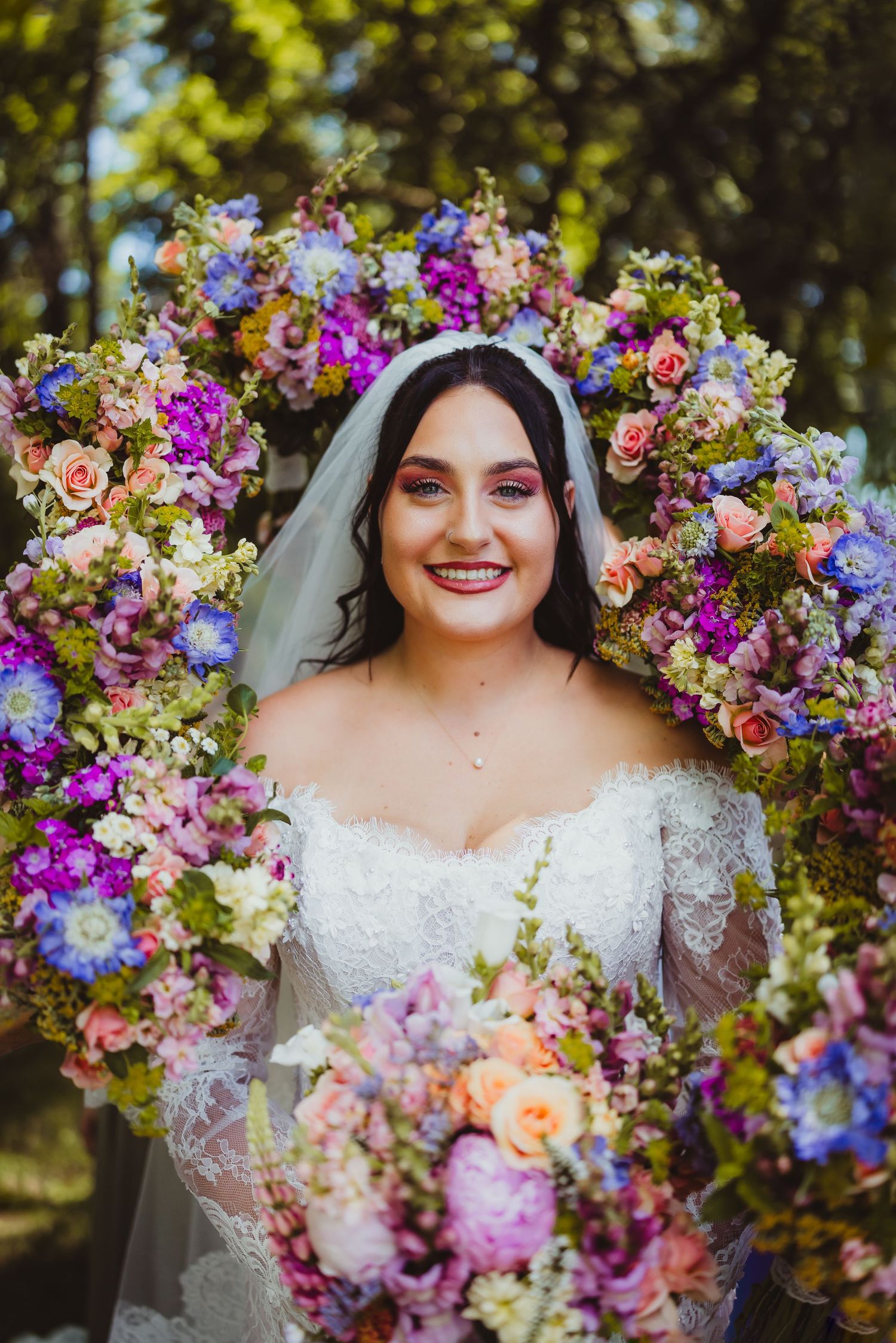 Smiling bride in white lace gown holding bouquet, surrounded by vibrant purple and pink floral wreath outdoors.