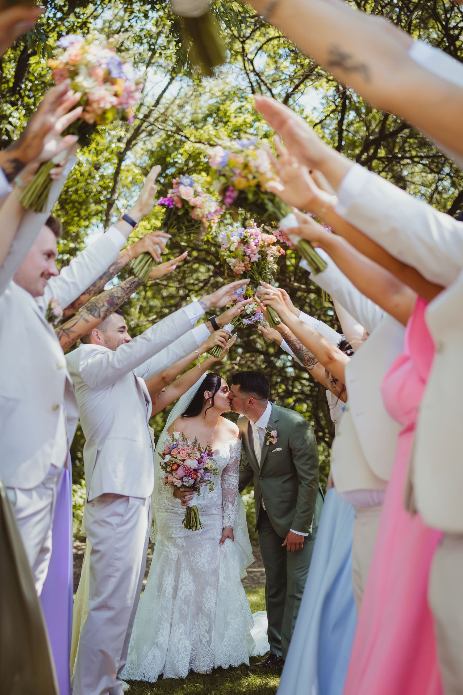 Bride and groom kiss under an arch of raised bouquets held by wedding party guests outdoors.