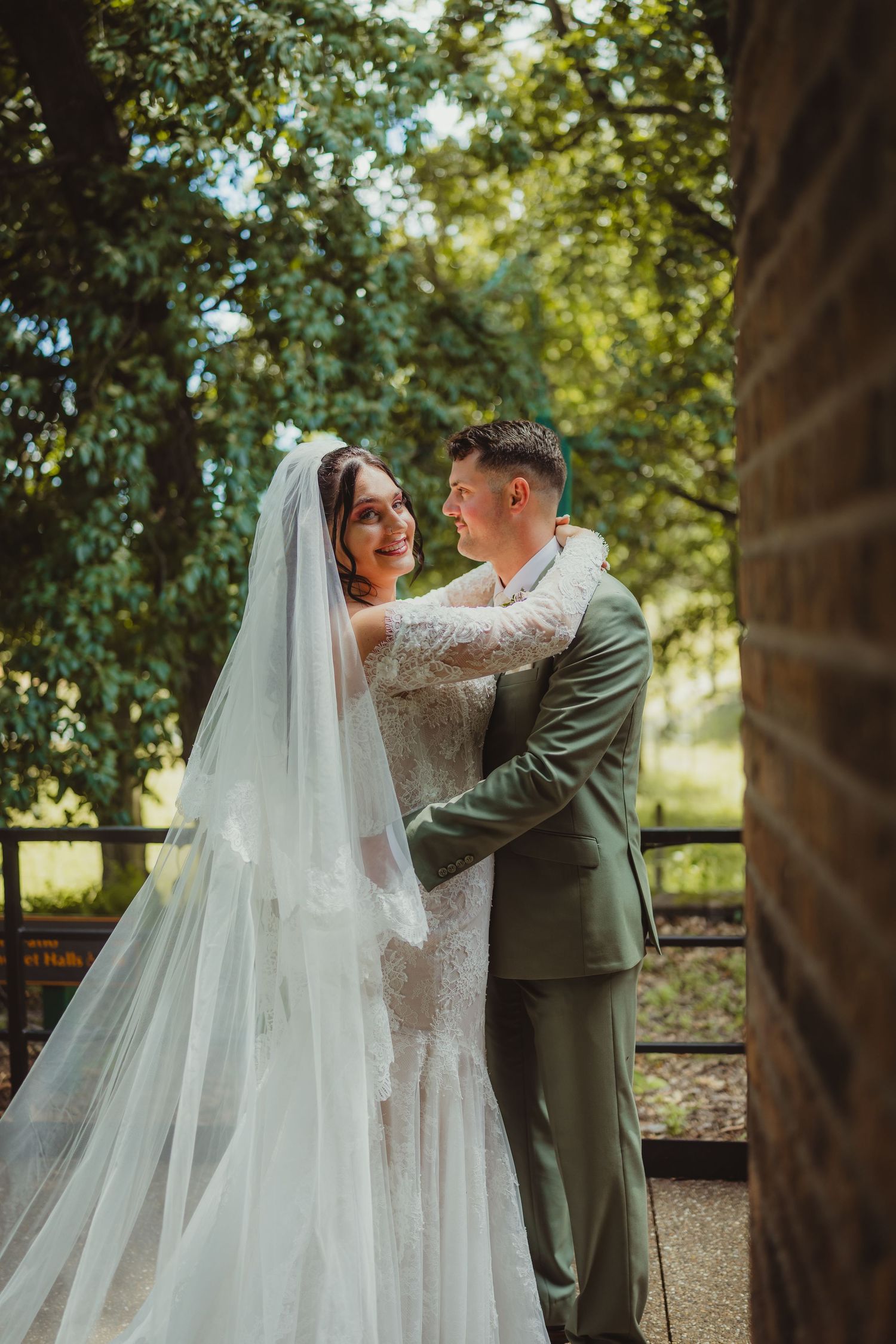 Bride and groom embrace outdoors, she wears a lace gown with veil, he wears a sage green suit, surrounded by lush trees.