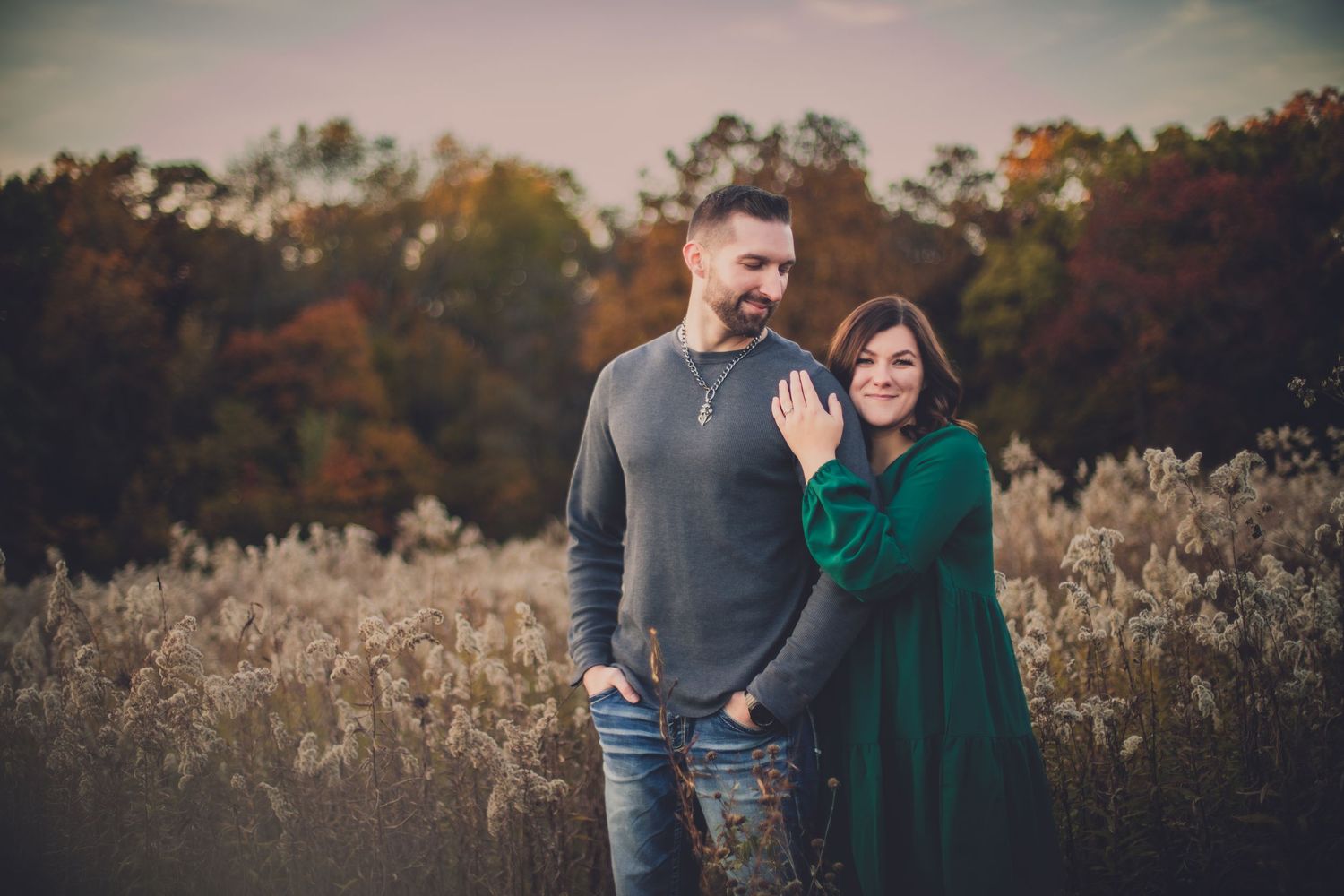 Couple in casual wear pose together in field of autumn grasses during sunset.
