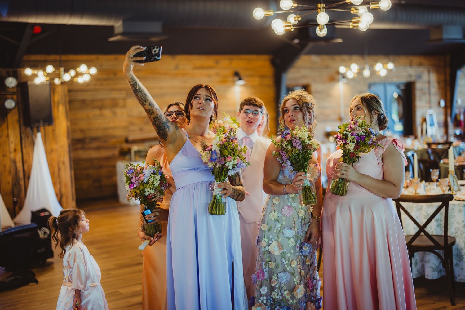 Bridesmaids in pastel gowns taking a selfie while holding colorful bouquets at a rustic wedding venue.