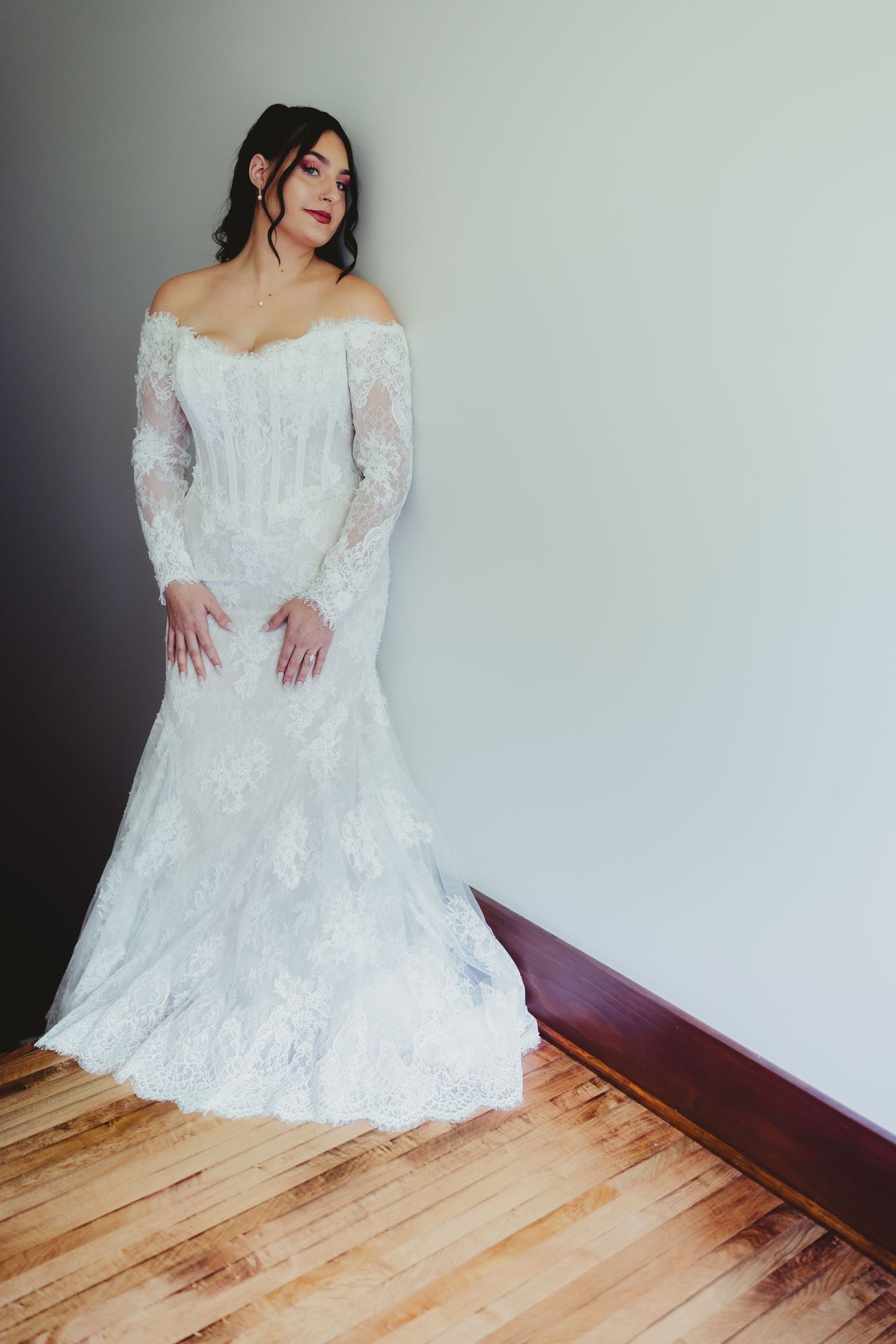 Bride in elegant off-shoulder long-sleeve white lace wedding gown posing against a light gray wall indoors.