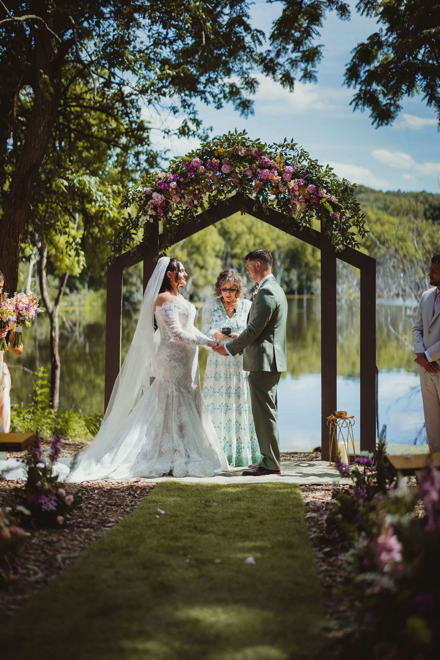 Bride and groom exchange vows under a floral hexagonal arch by a lake, officiated by a woman in a green dress.