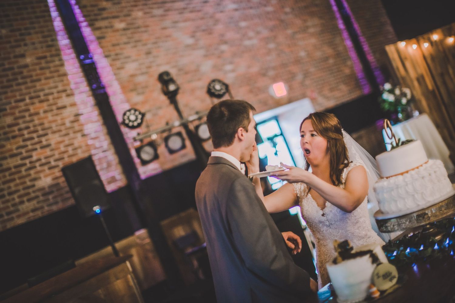 A couple playfully feeds each other cake during their wedding reception in an industrial-style venue.