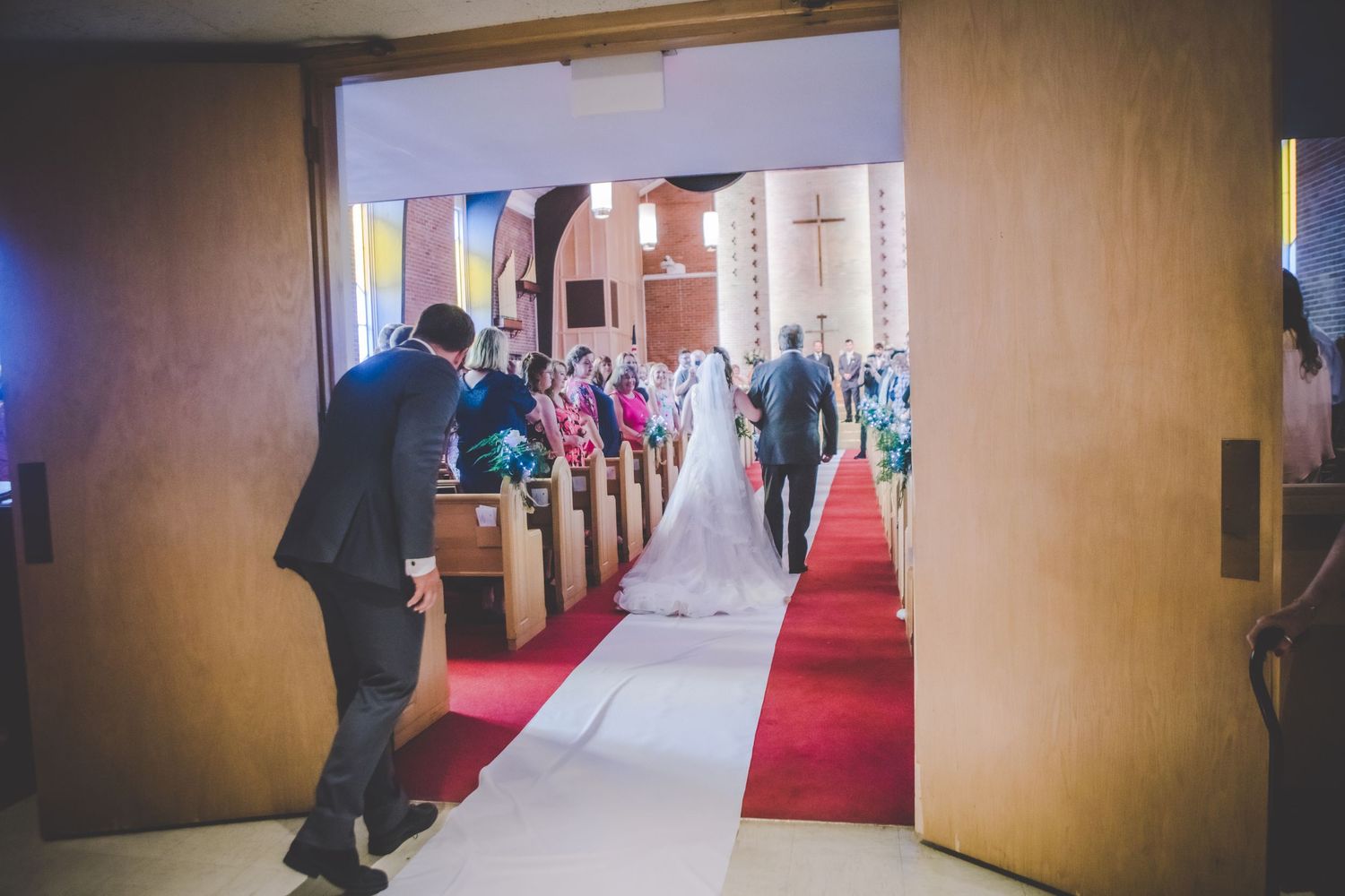 View through doorway of a wedding ceremony with guests seated in wooden pews along a red carpet aisle.