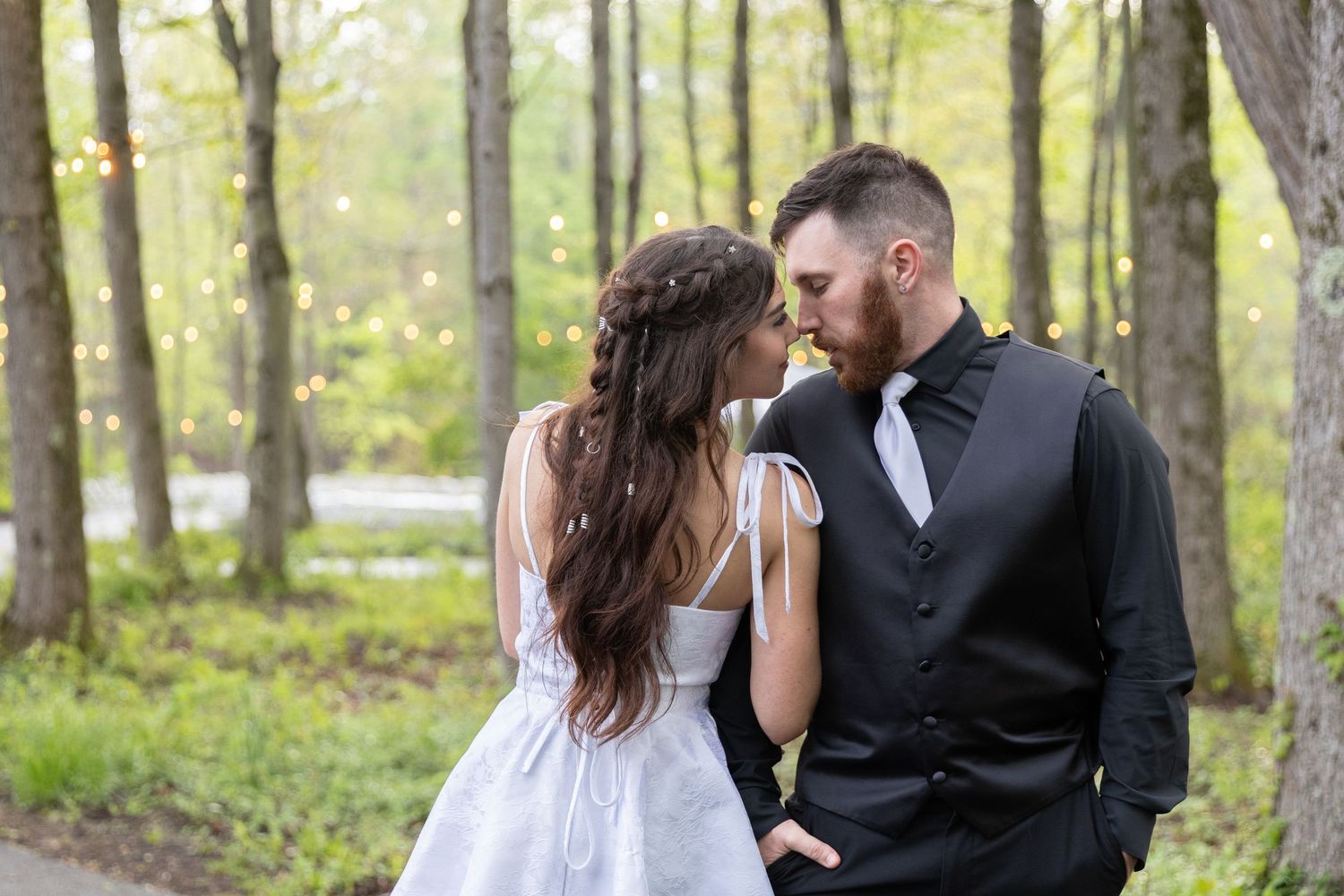 A bride and groom share a romantic moment in a lush green forest, dressed in wedding attire.