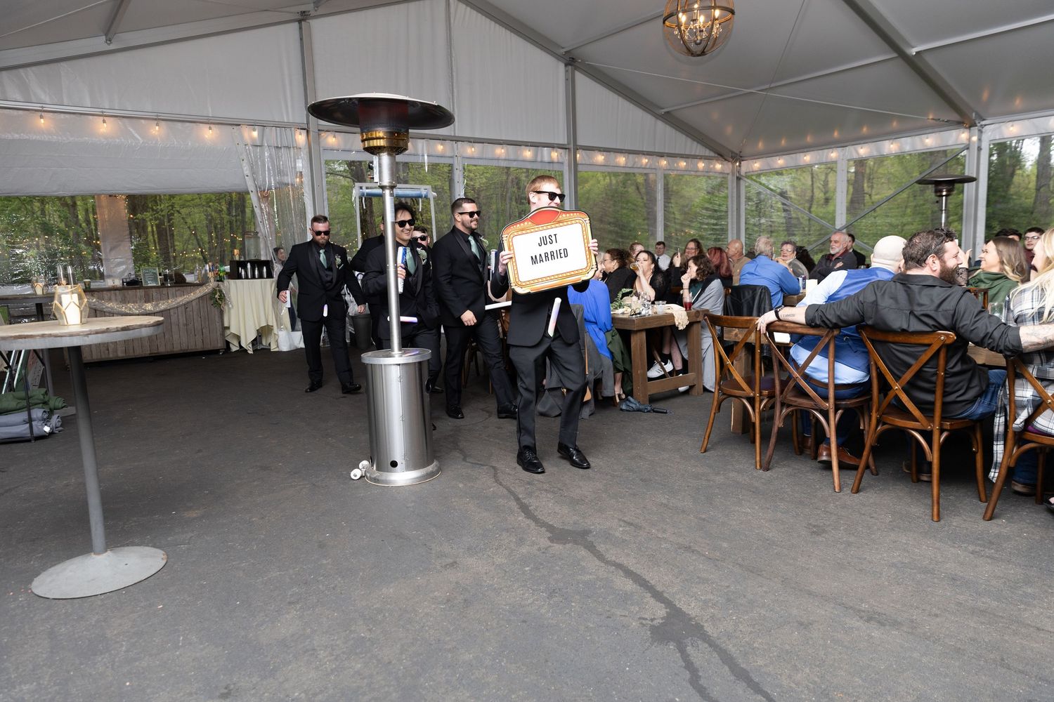 Outdoor event under a tent with guests seated at tables, servers in black attire, and patio heaters on a paved area.