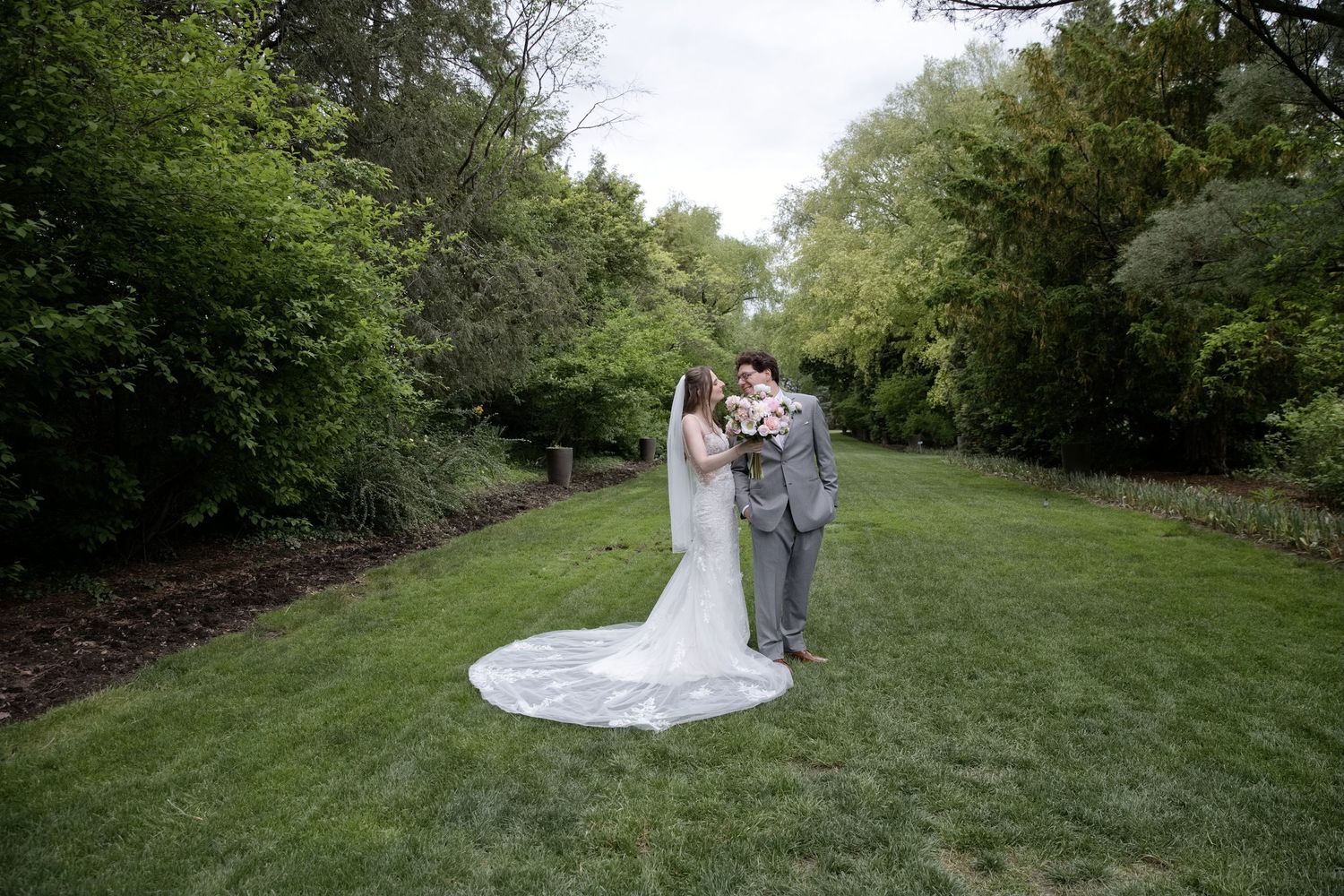 Bride and groom share a kiss on lush green lawn surrounded by trees, bride's long veil trailing behind her.