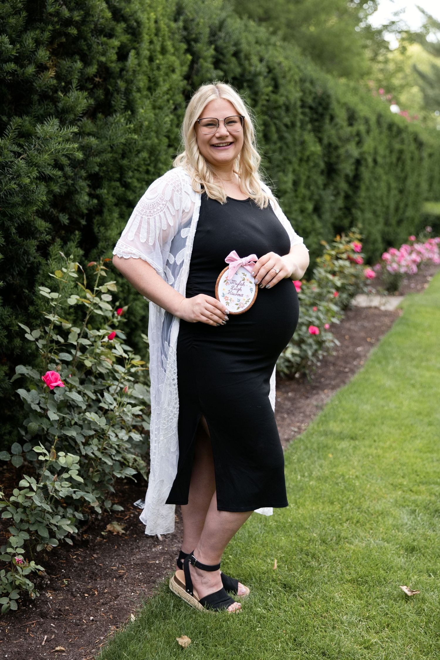 Pregnant woman in black dress and white lace cardigan holds baby girl sign in garden setting.