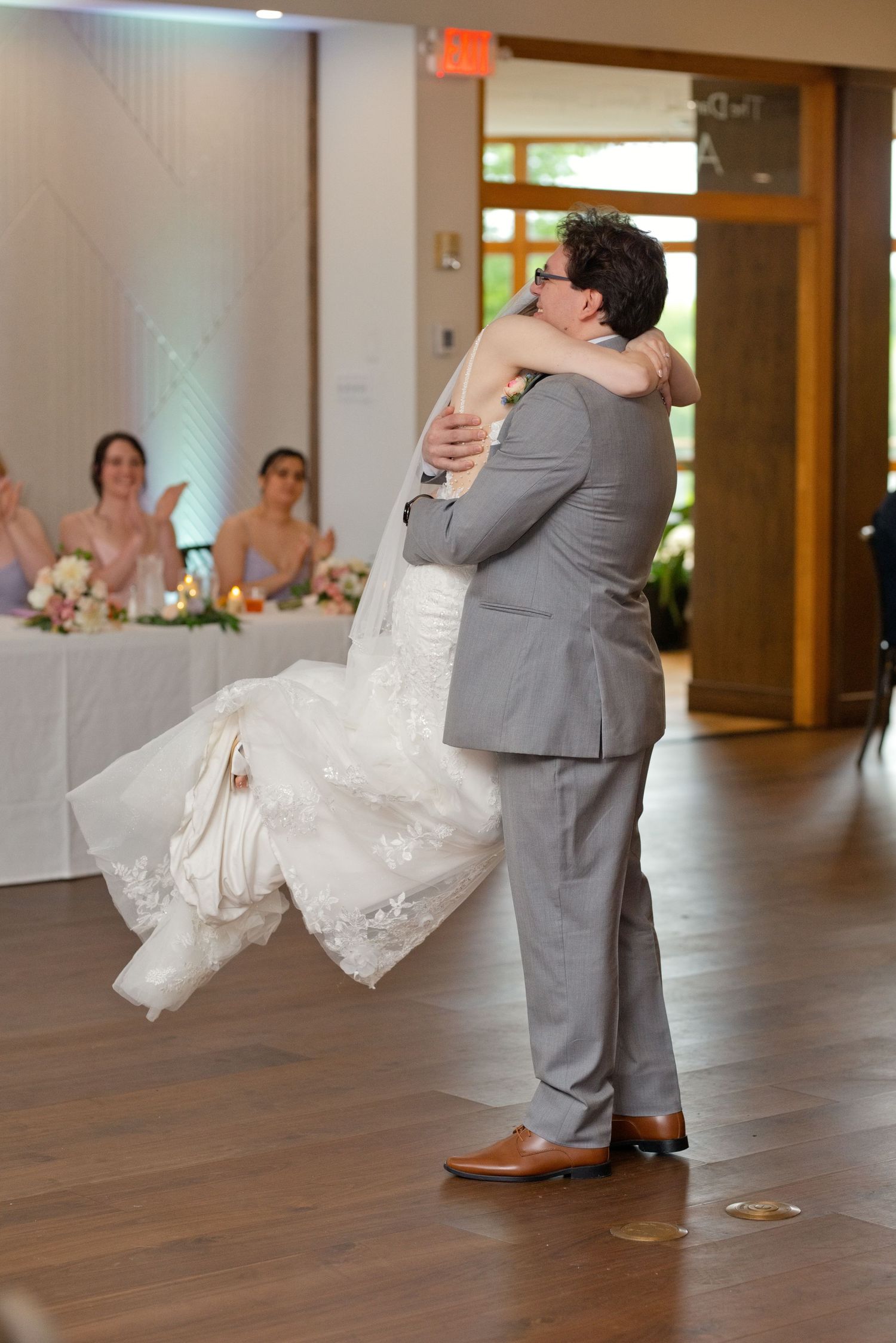 Groom in gray suit lifts bride in white lace wedding dress during first dance at elegant reception venue.