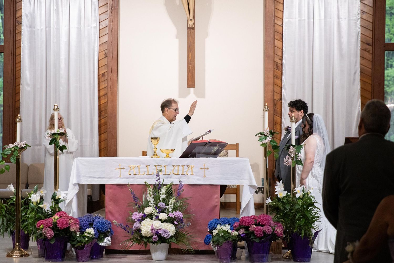 Priest officiating a wedding ceremony at church altar with bride and groom, surrounded by floral arrangements.