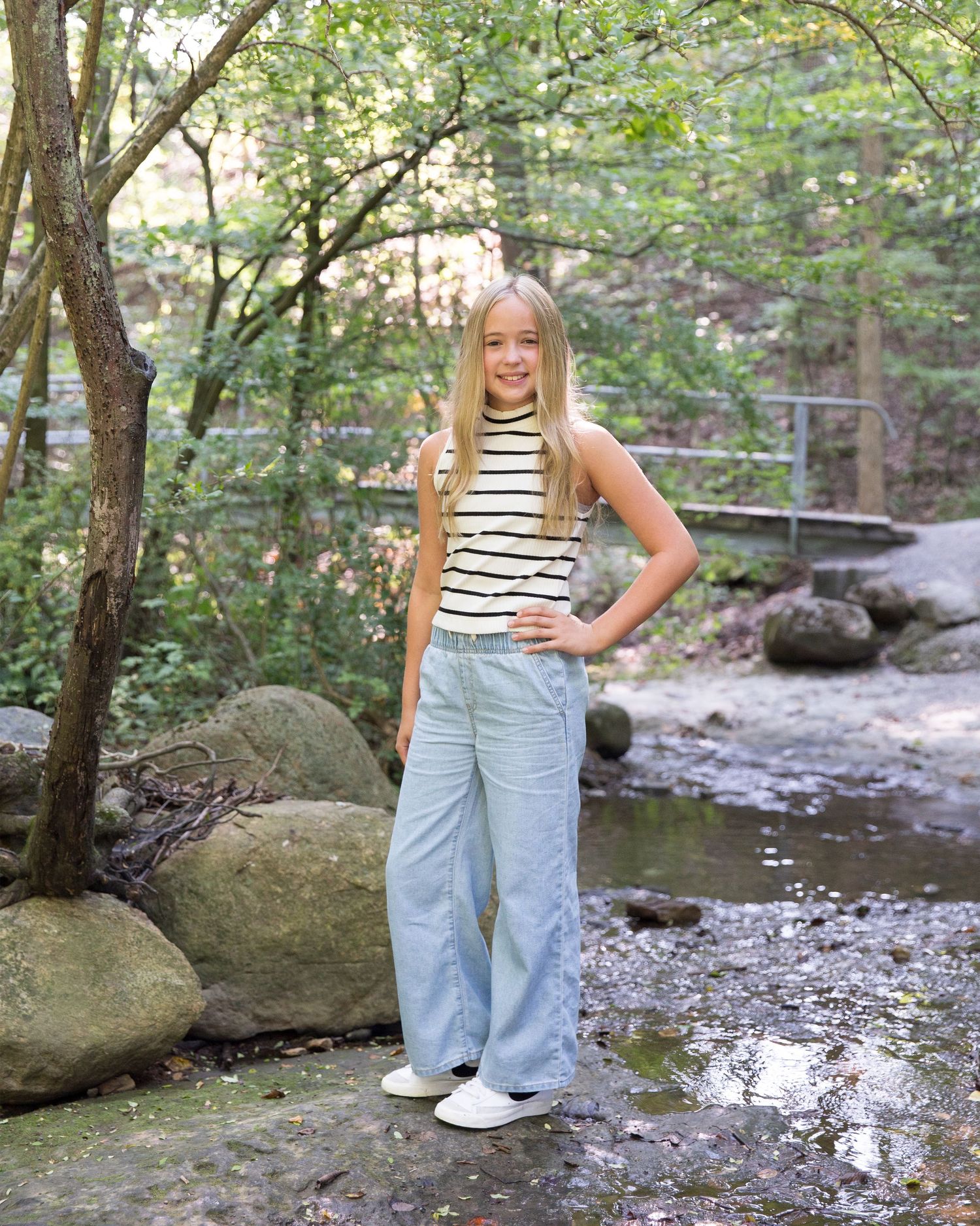 Person in striped shirt and overalls standing on rocks by woodland stream.