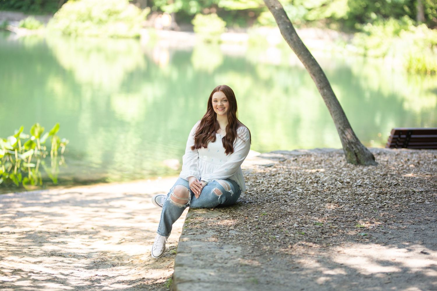Young woman in a white shirt sitting, smiling, with trees and sunlight in the background.